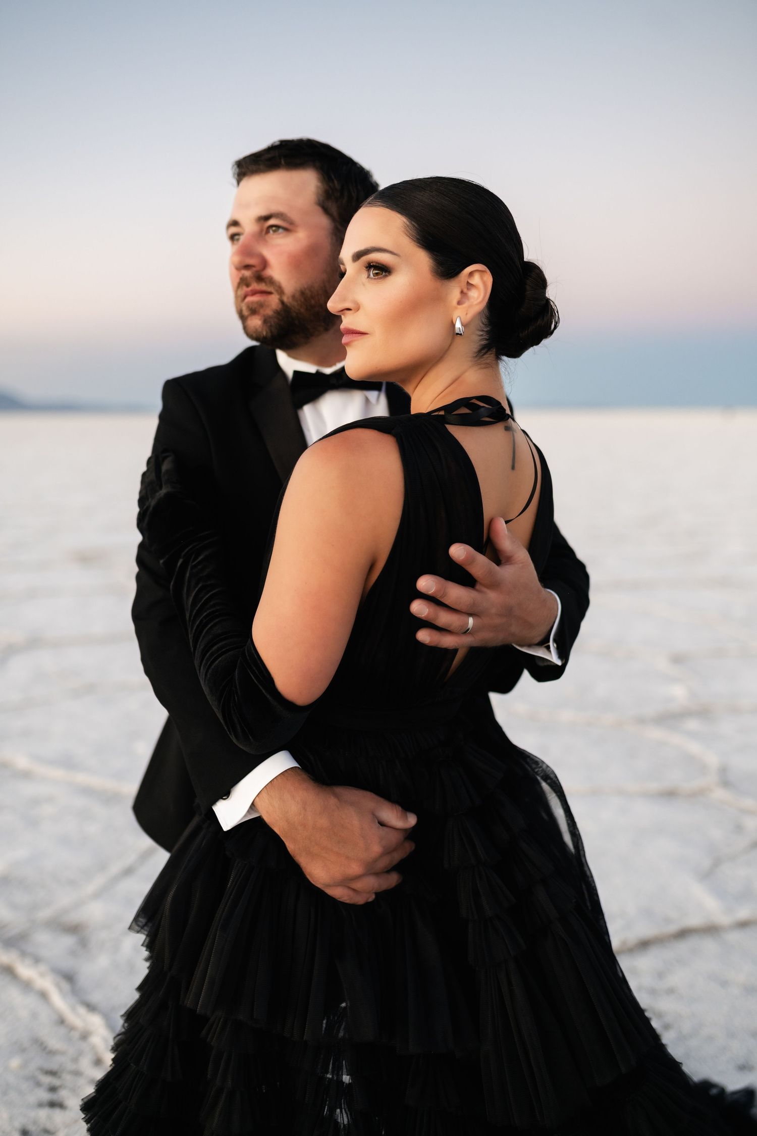 Utah Salt Flats Photographer. A romantic couple in black formal wear embraces while standing on salt flats during sunset.