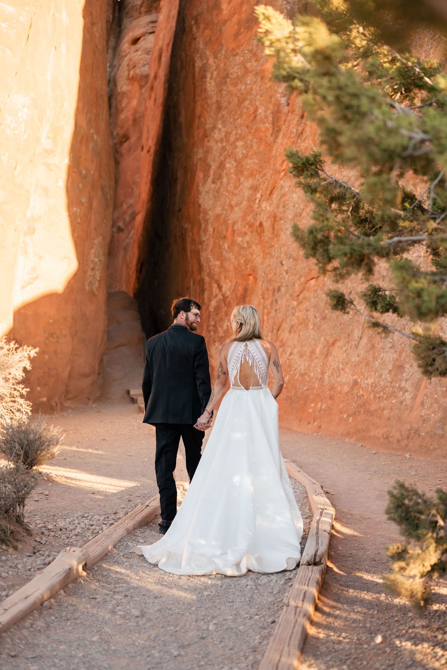 A couple in wedding attire walks along a scenic red rock canyon path in soft sunlight.