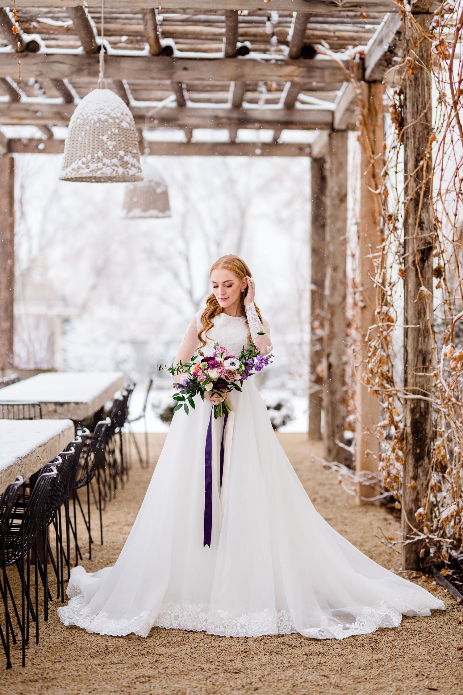 Bride in white lace ballgown poses with purple bouquet in rustic barn venue with wooden beams at The Wild Oak.