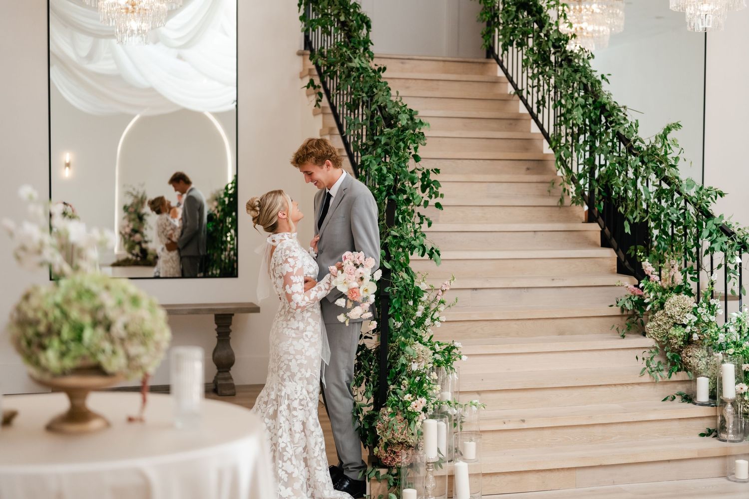 Elegant wedding couple embraces on grand staircase adorned with lush greenery and crystal chandeliers at Moments on Main in Payson