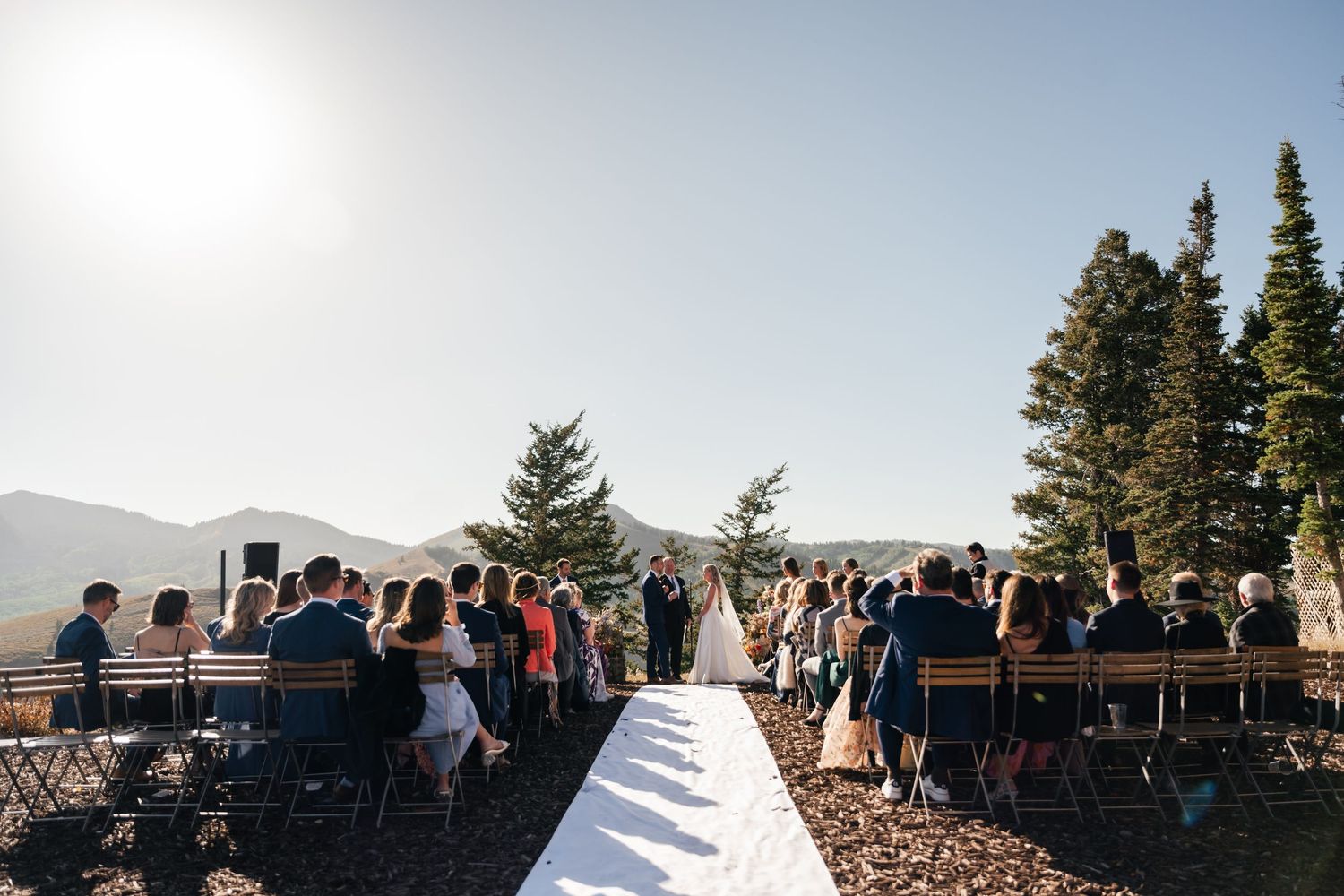 Mountain wedding ceremony with guests gathered on a scenic overlook surrounded by evergreen trees at sunset.