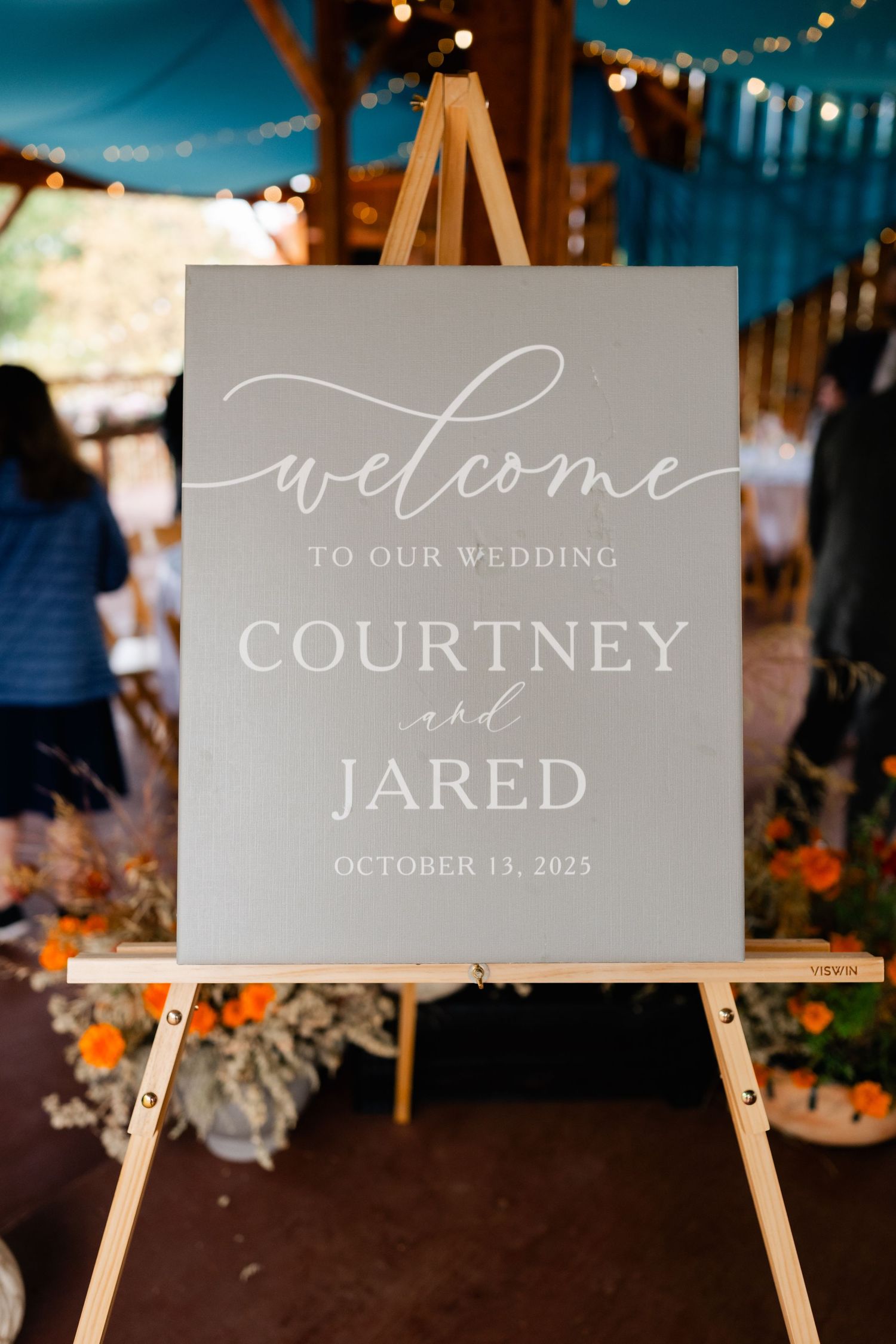Gray welcome signs on wooden easels displaying wedding details with string lights in the background.