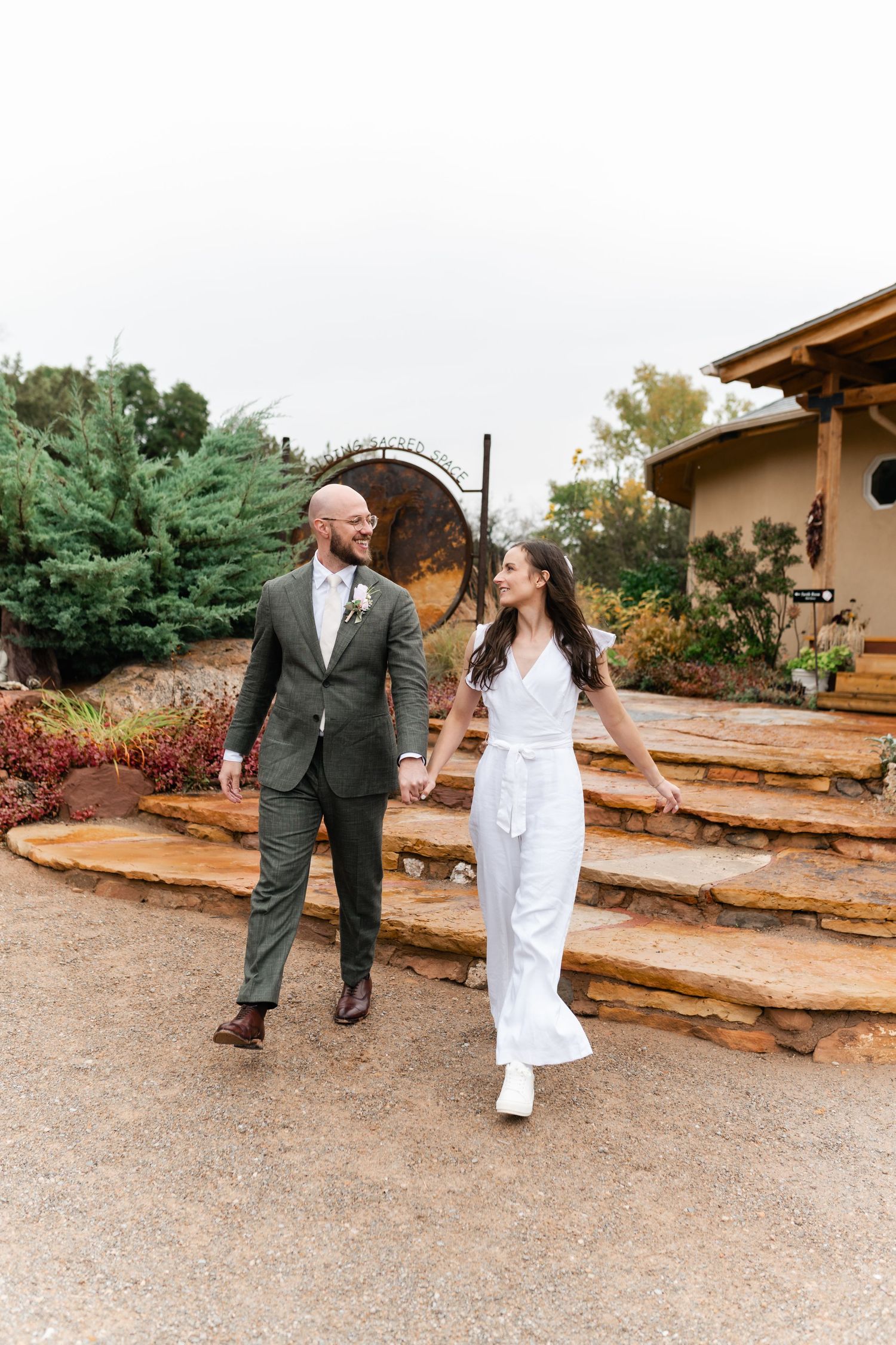 A couple walks hand in hand along a rustic path surrounded by greenery.