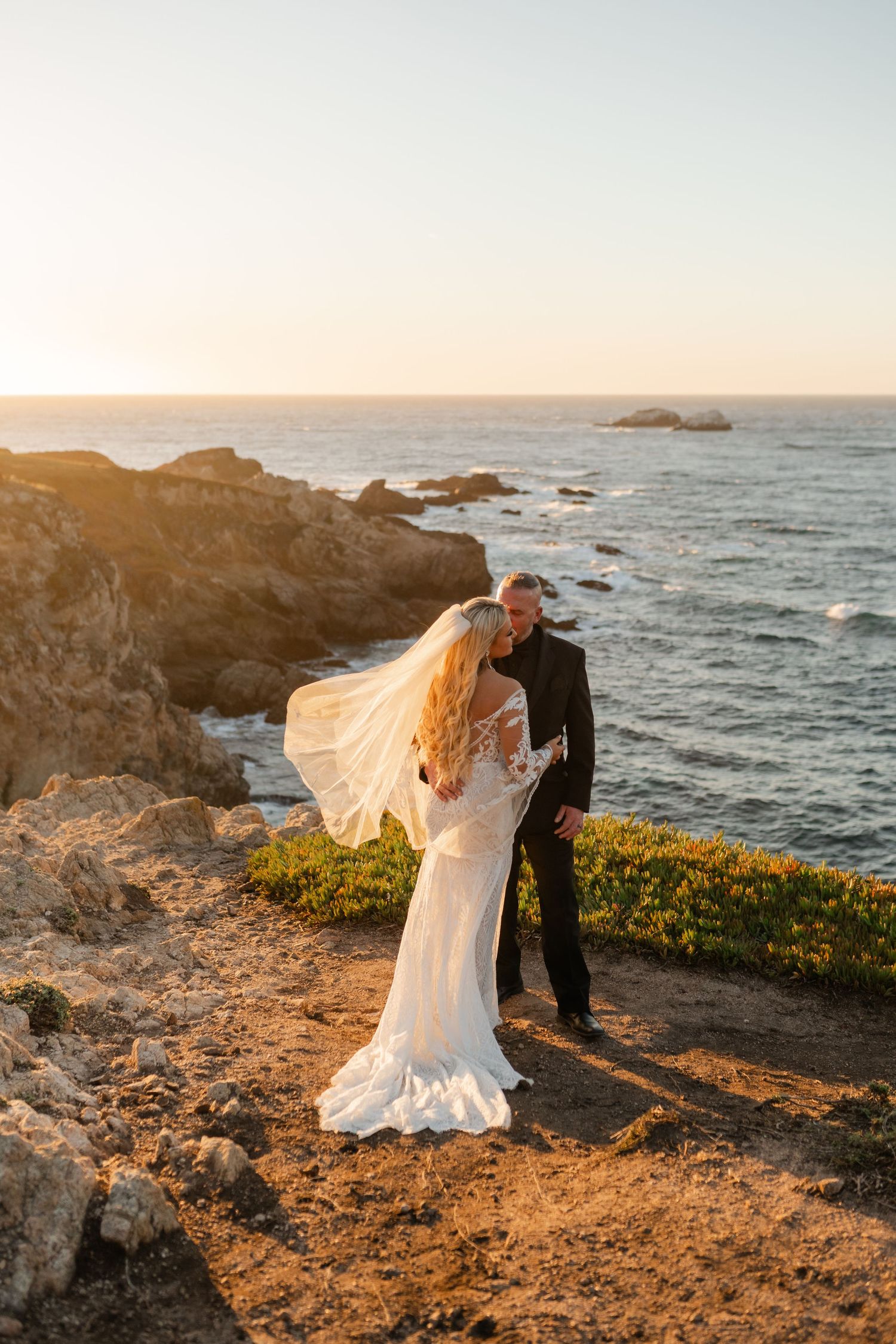 Couple in wedding attire embracing on coastal cliffs at sunset with ocean waves crashing below.