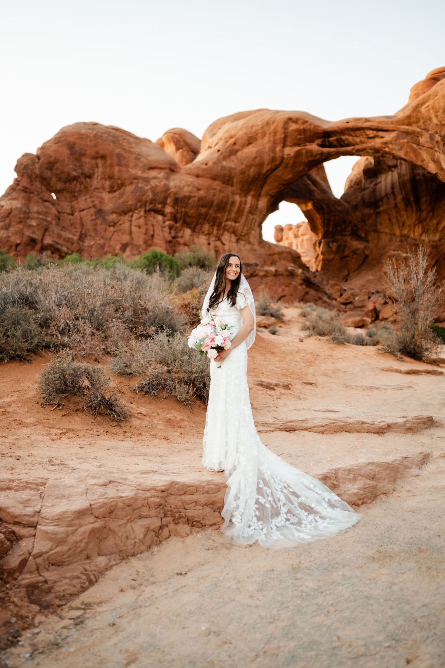 A white wedding dress flows elegantly against the red rock formations of a desert landscape at sunset.