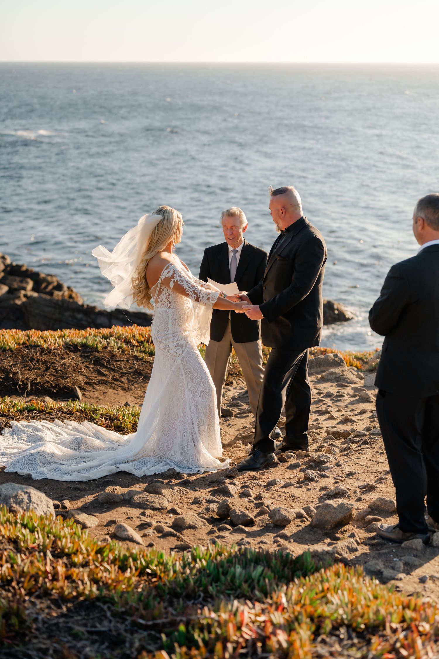An intimate beach wedding ceremony takes place on a scenic oceanside cliff at sunset.