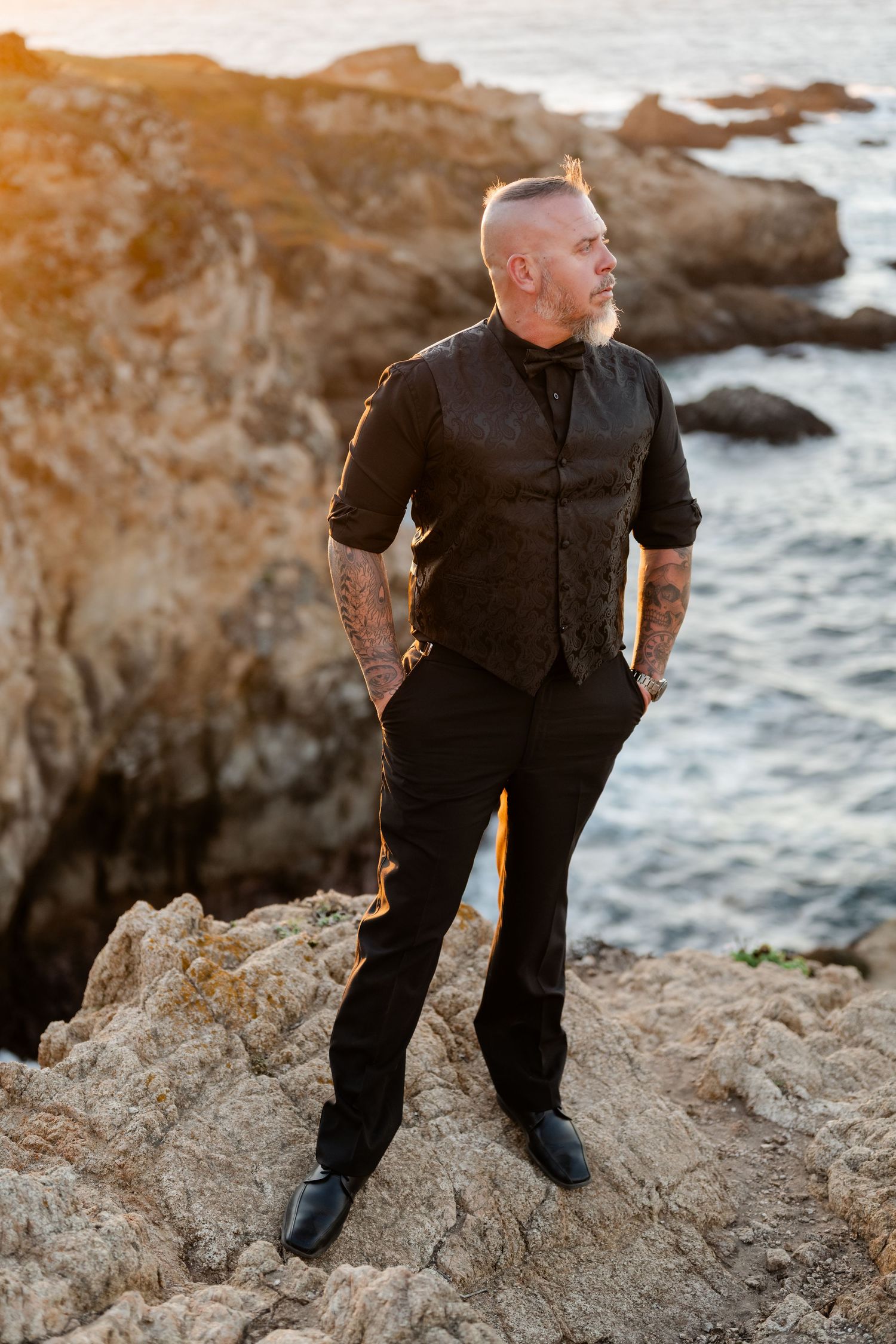 Stylish figure in all black attire poses confidently on rocky coastal cliff during golden hour.
