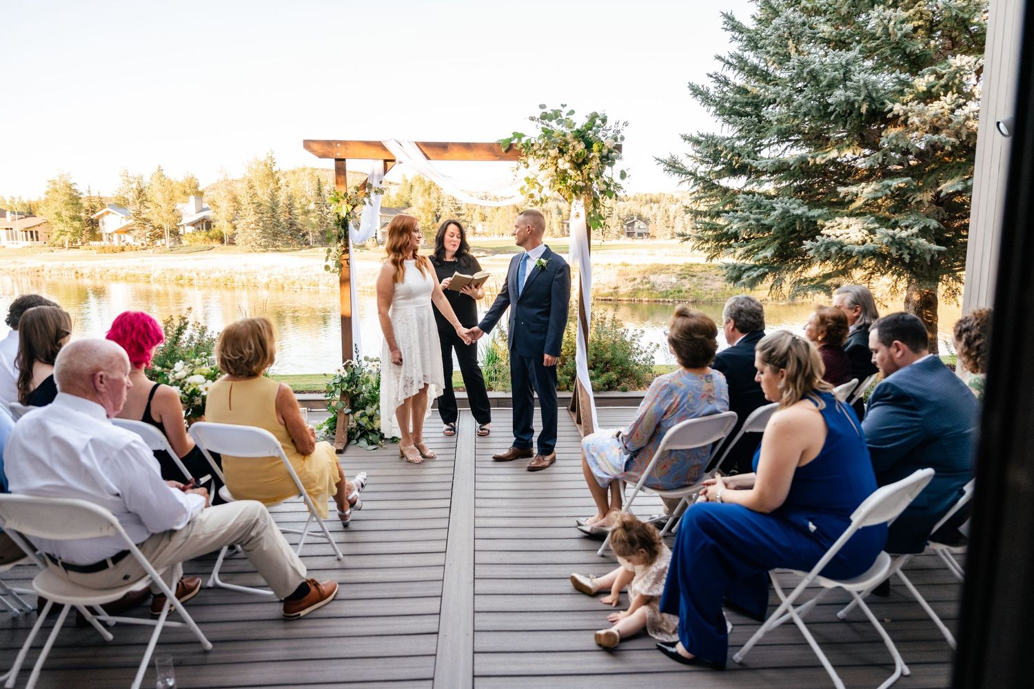 An intimate outdoor wedding ceremony on a deck overlooking a lake with wooden arch and white draping.