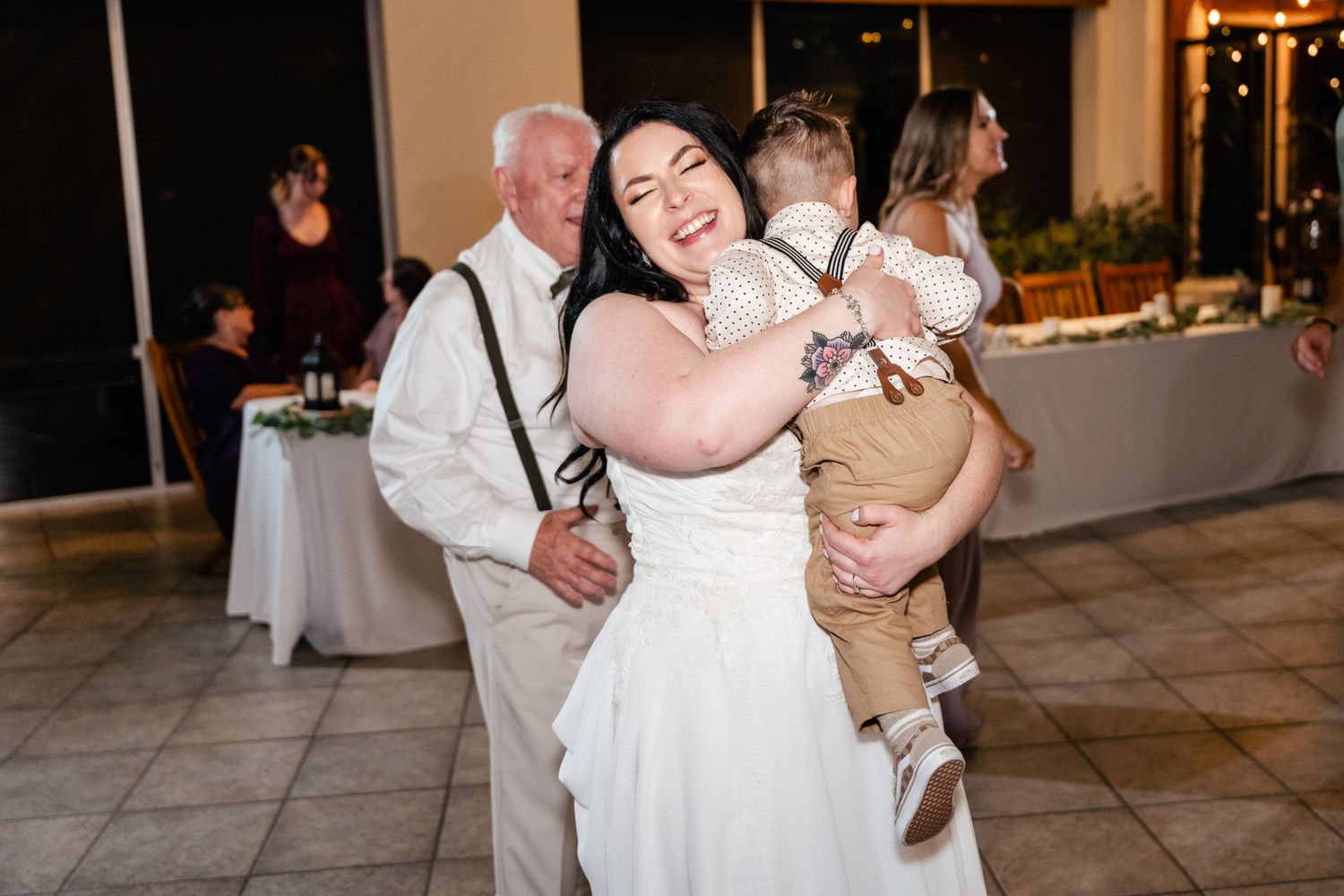 A bride in a white wedding dress dances while holding a small child at an indoor reception venue.
