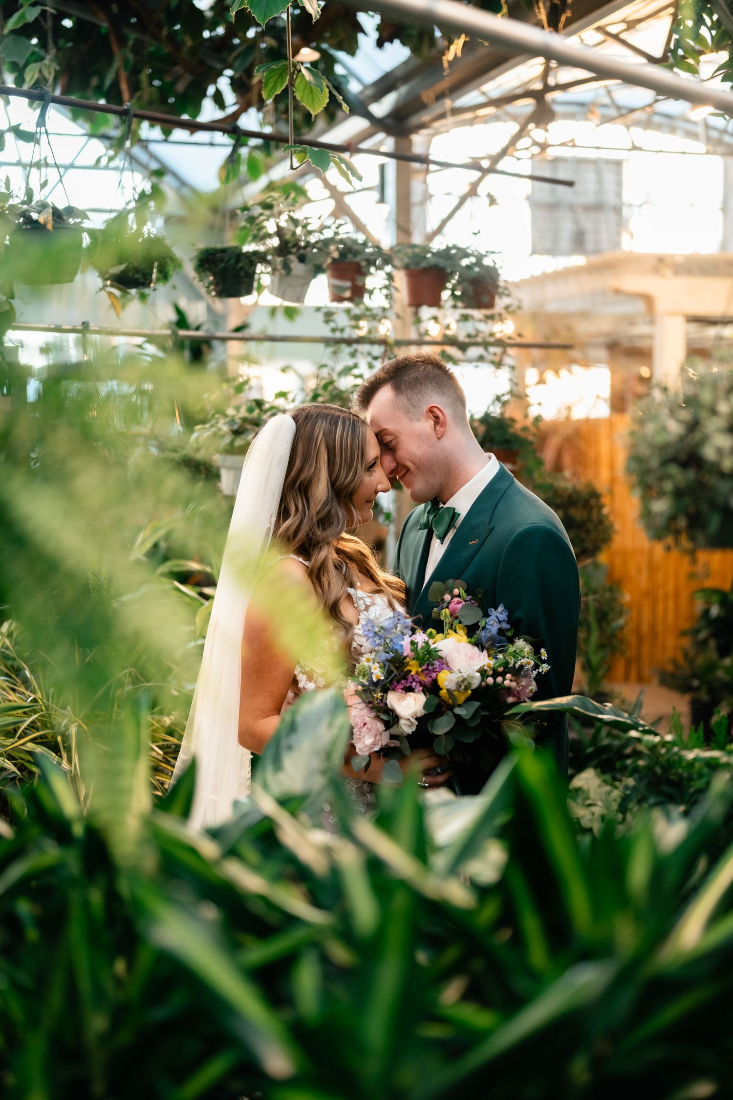A romantic wedding moment captured in a lush greenhouse setting with vibrant florals and greenery framing the scene.