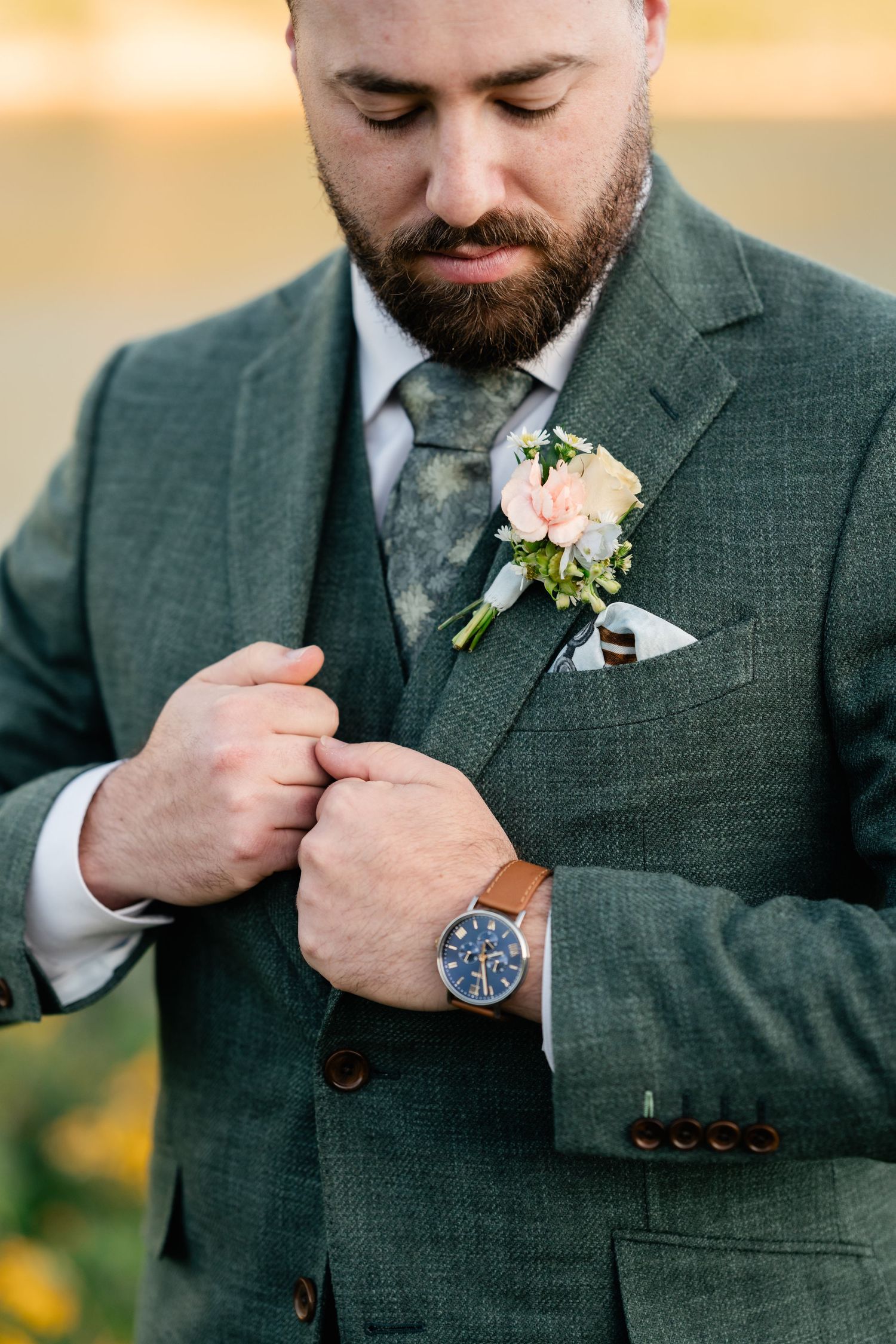 Close-up of groom in forest green suit with floral boutonniere adjusting watch and lapel.