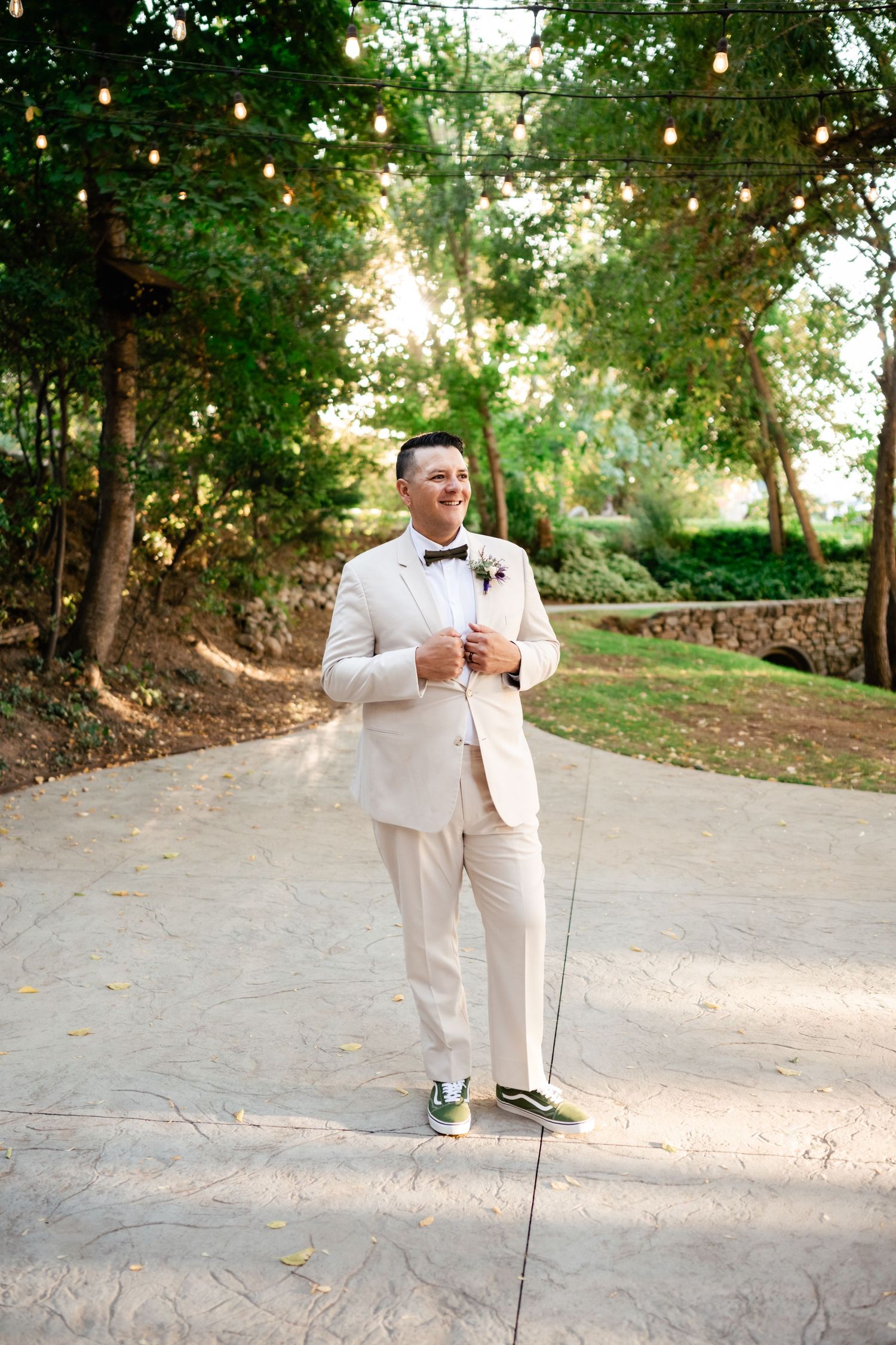 A groom in a cream colored suit and bowtie stands on a garden path with string lights overhead.