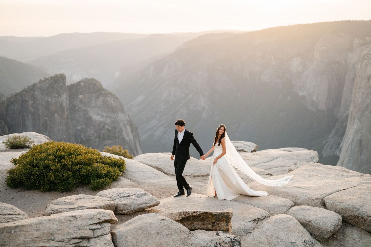 Bride and groom hold hands on rocky Yosemite cliff at sunset with dramatic mountain valley views.