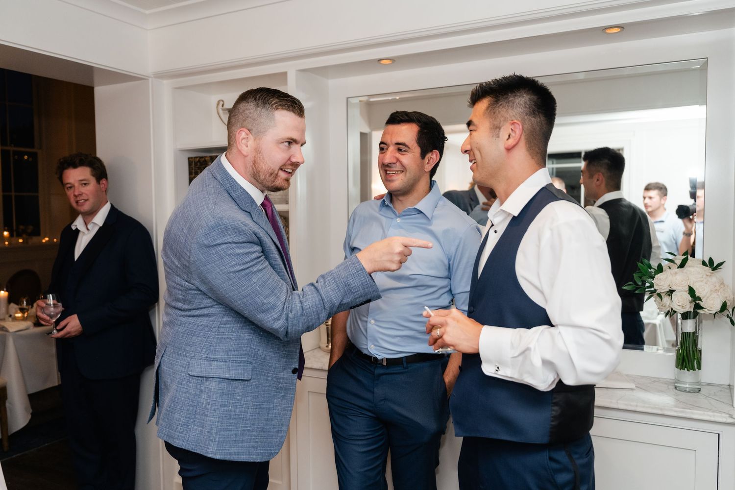 Three men in suits laughing and chatting during wedding reception cocktail hour indoors.