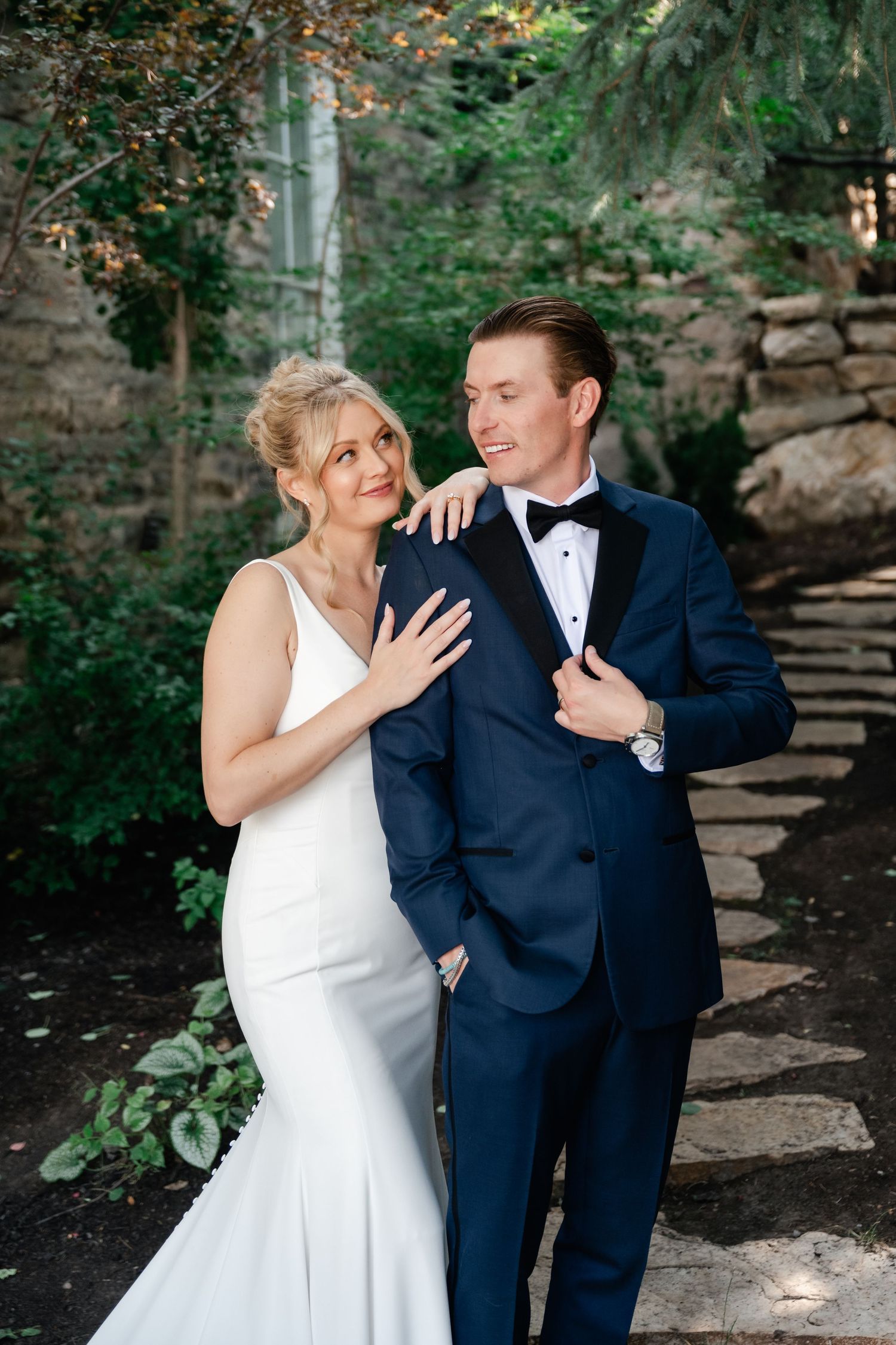 Bride in white gown and groom in navy tuxedo embrace outdoors near stone wall and lush greenery during their Park City micro wedding.
