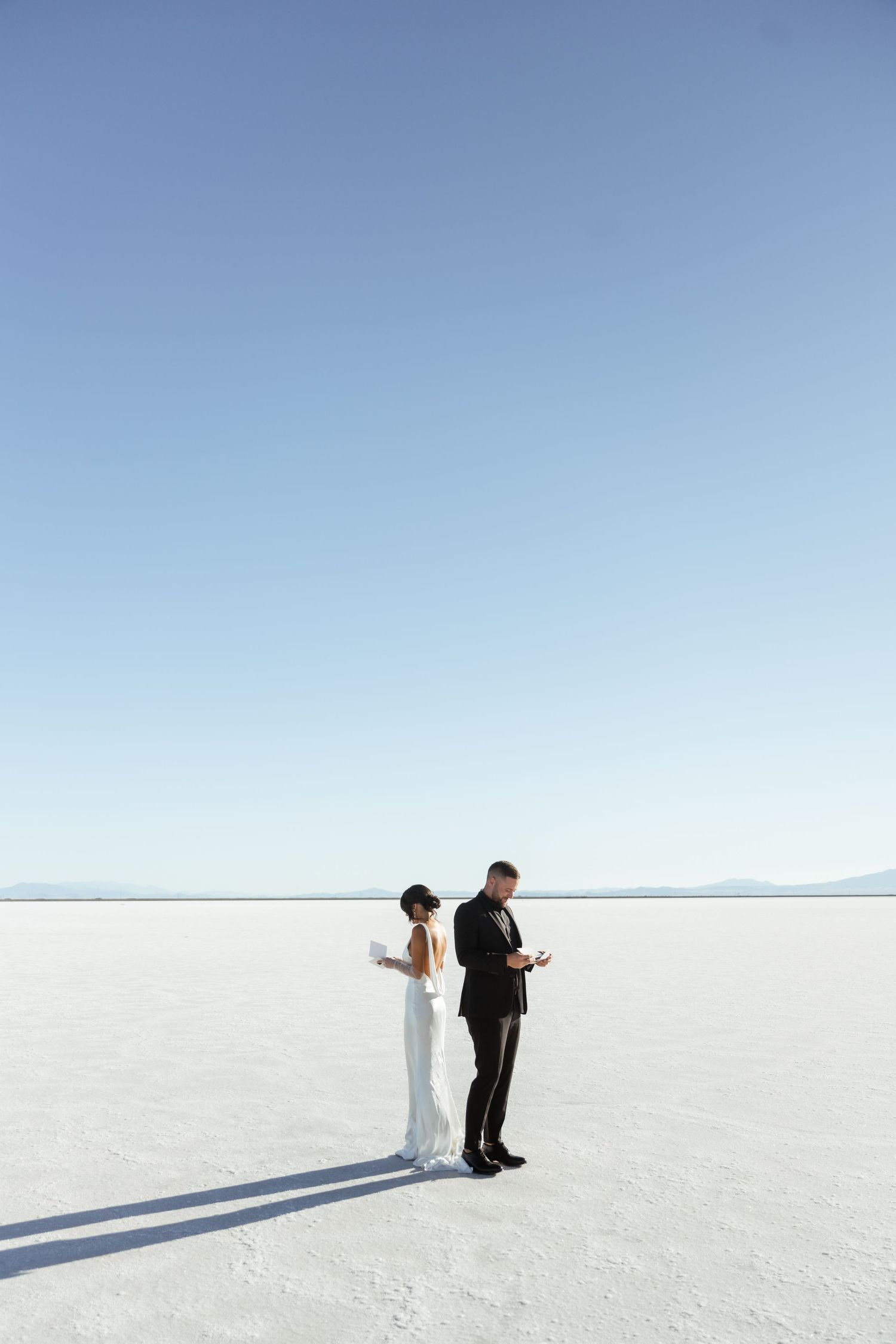 Wedding couple creating dramatic silhouettes against bright white salt flats under blue sky.