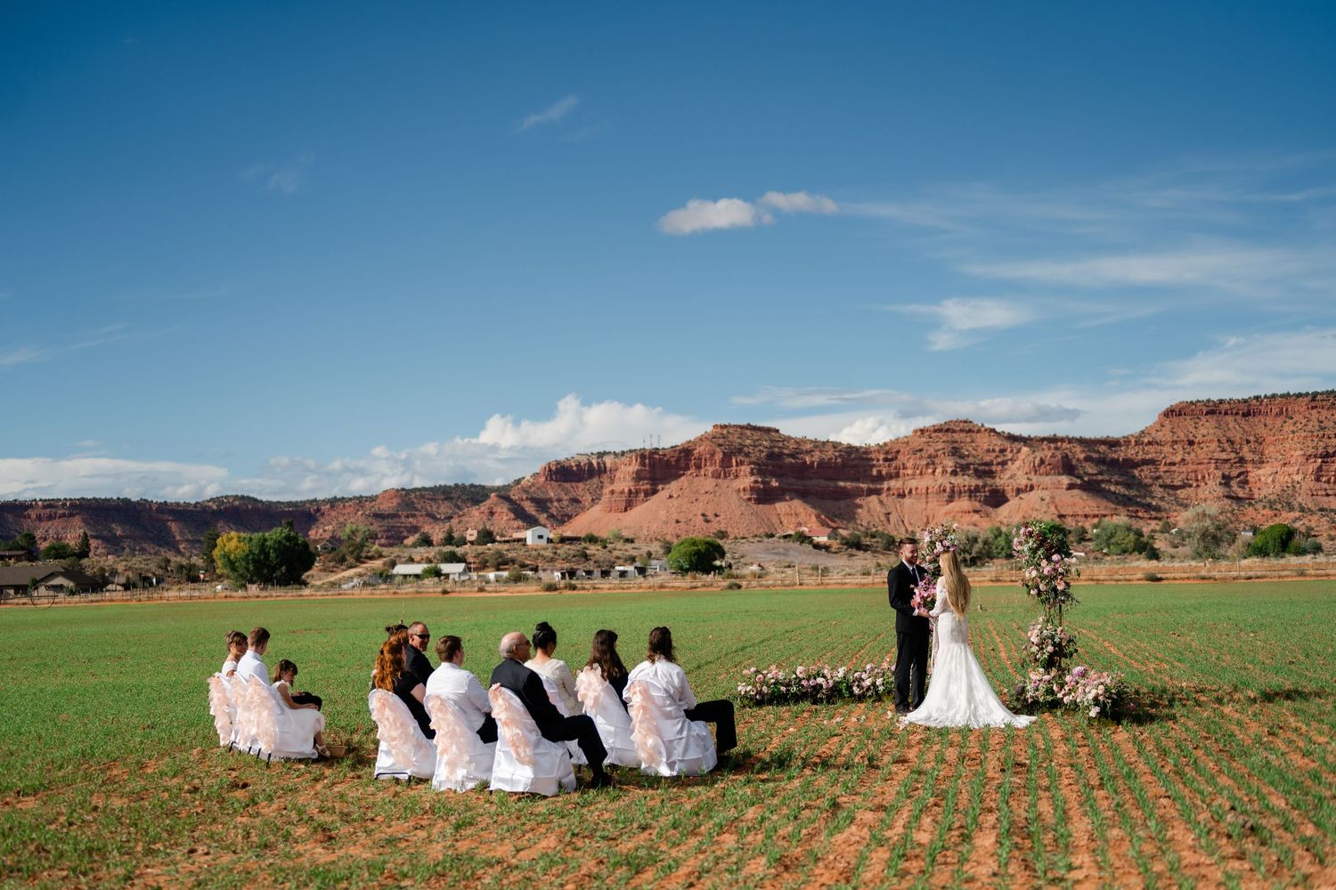 Wedding ceremony taking place on a green field against dramatic red rock cliffs and blue sky in Utah. Southern Utah Wedding Photographer.