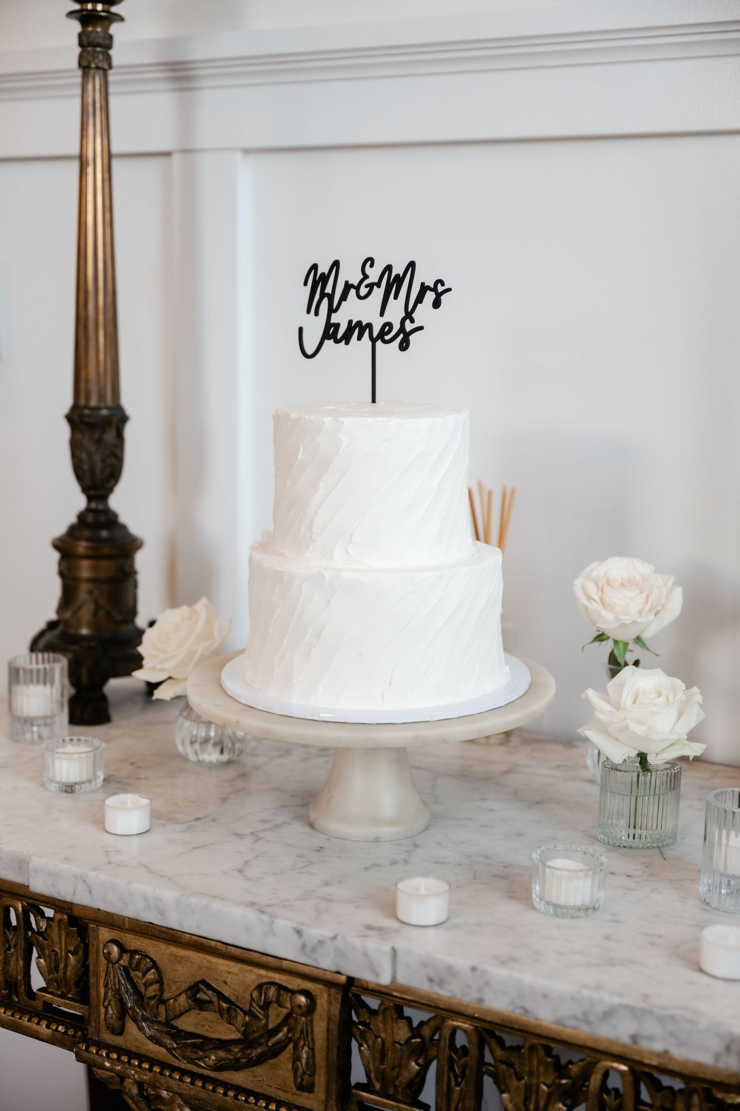 Elegant white wedding cake displayed on marble table with antique brass lamp and floral decorations.