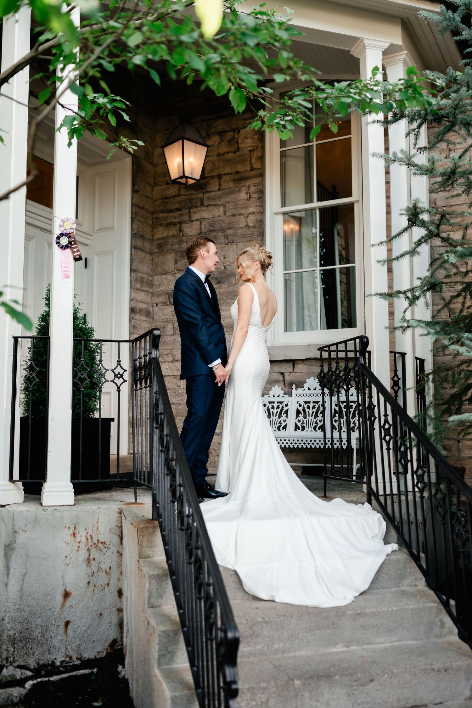 A bride and groom share an intimate moment on an elegant stone porch with black wrought iron railings and white columns.
