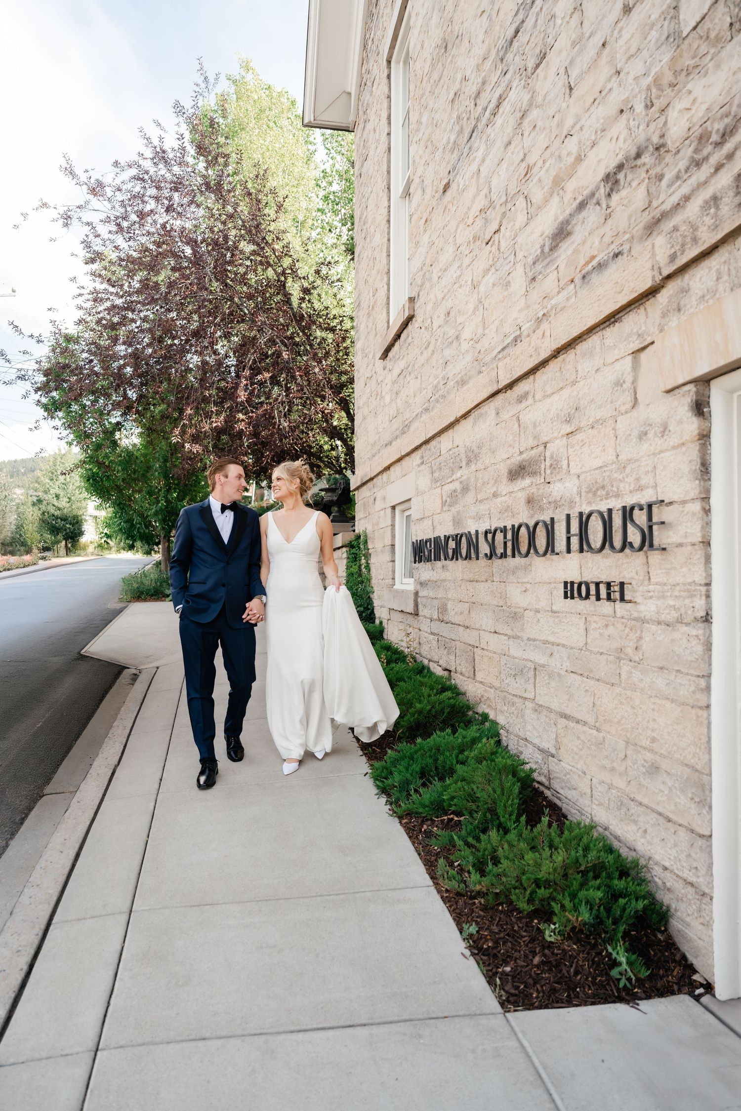 A couple in wedding attire walks along a stone building exterior with landscaping.