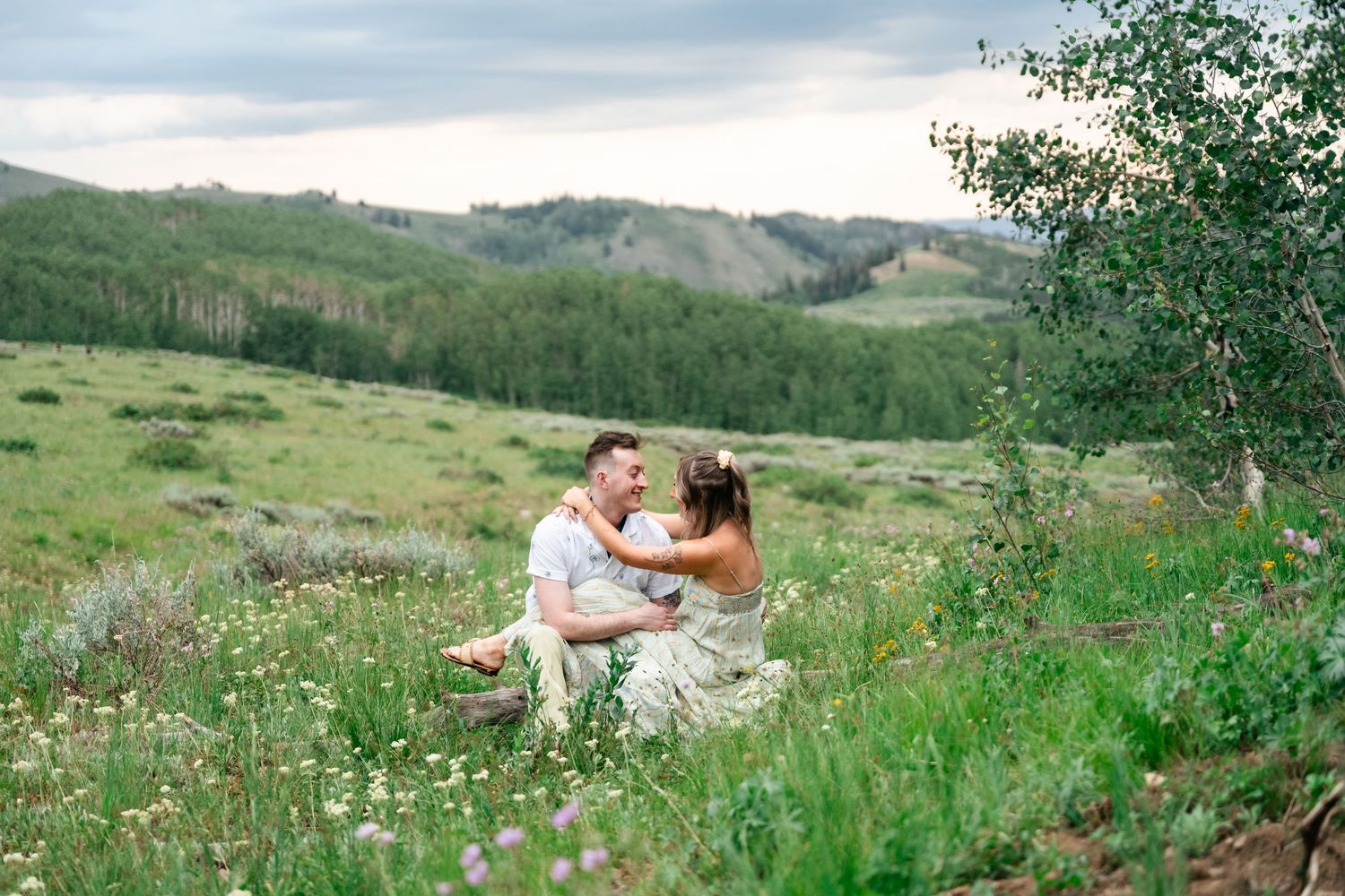 A couple relaxes in a scenic meadow surrounded by rolling green hills and wildflowers on a summer afternoon.
