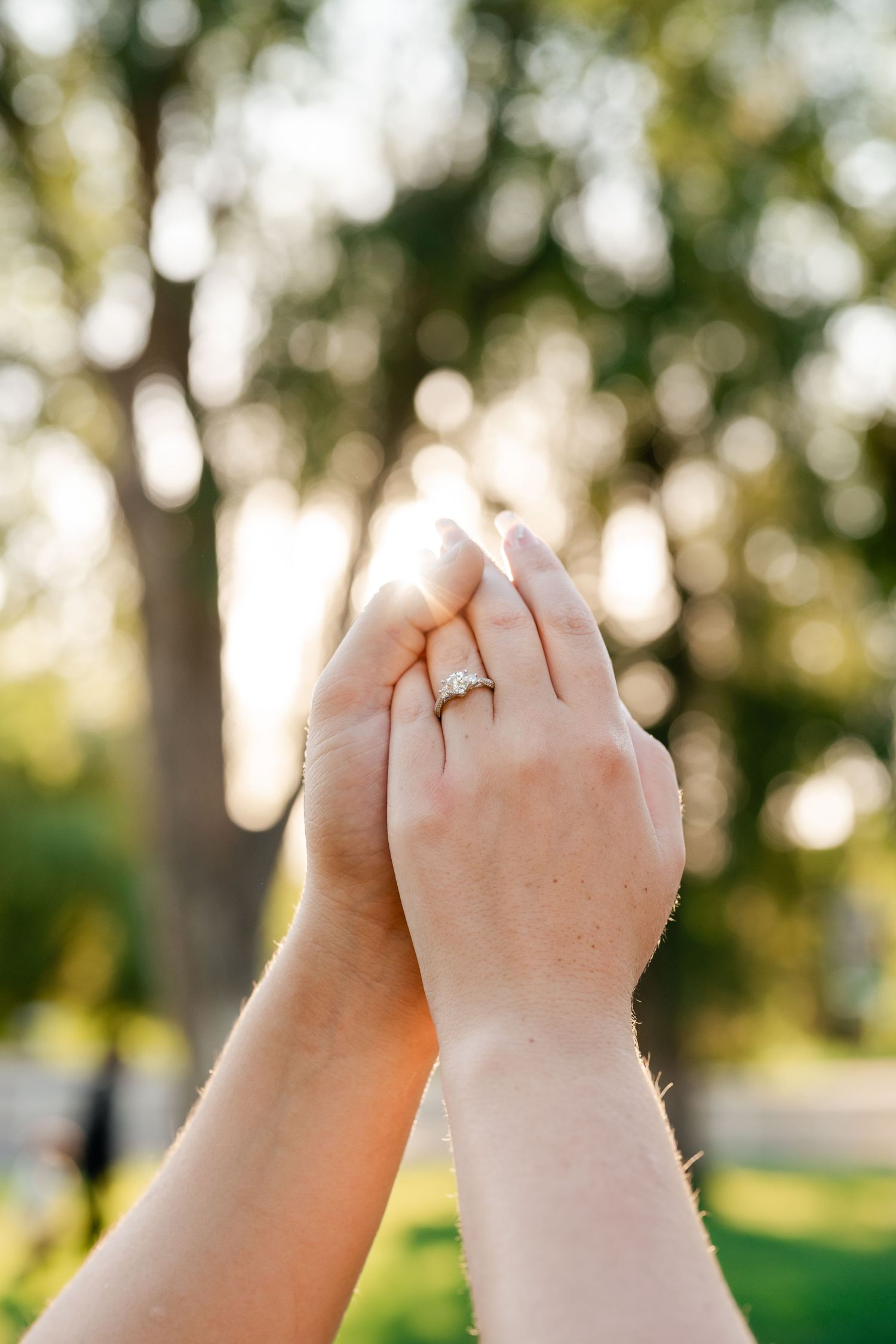 Two hands make pinky promise gestures against a blurred green outdoor background in sunlight. Ring detail photo by Utah Wedding Photographer