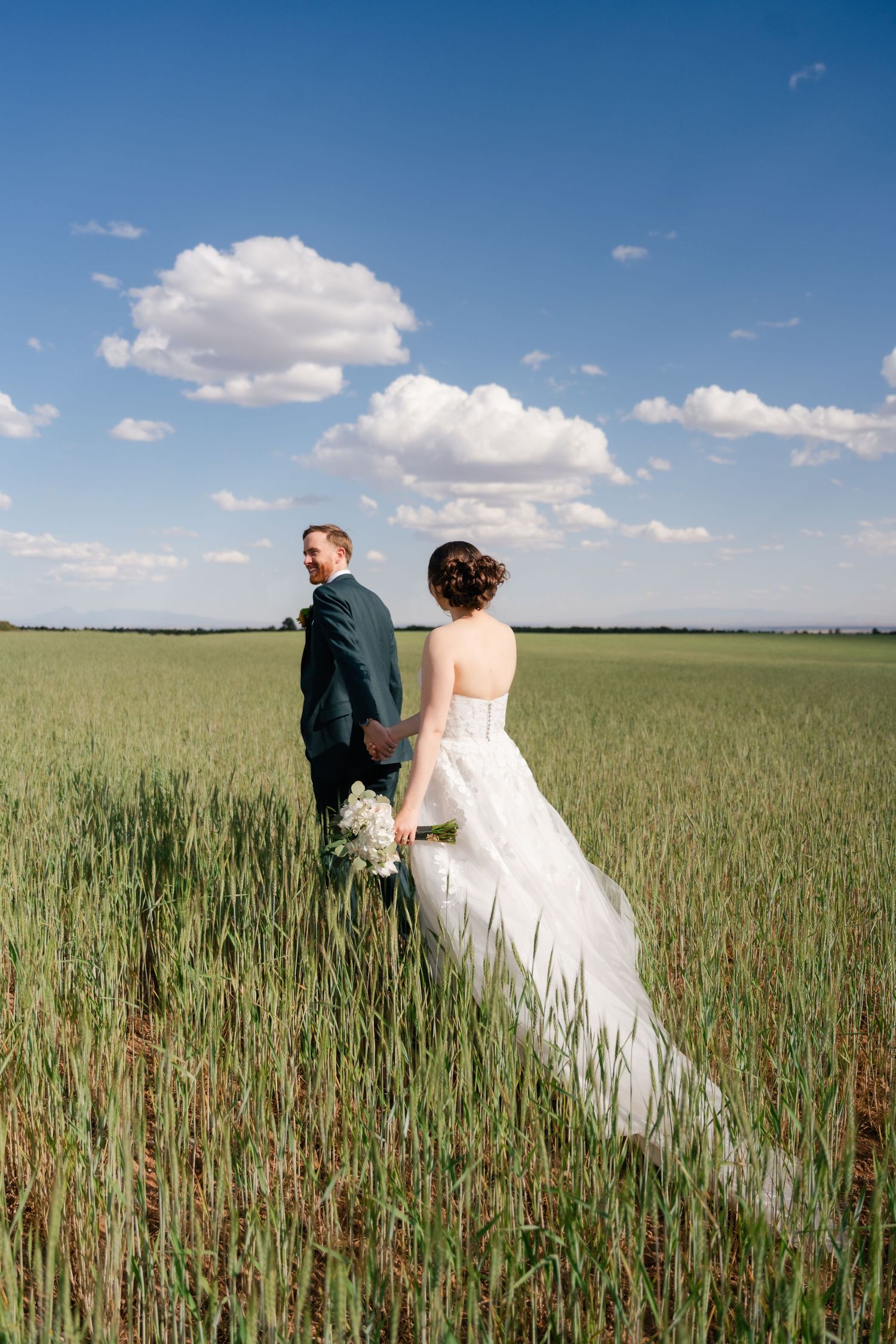 A couple in wedding attire walks through a golden wheat field under blue skies and white clouds.