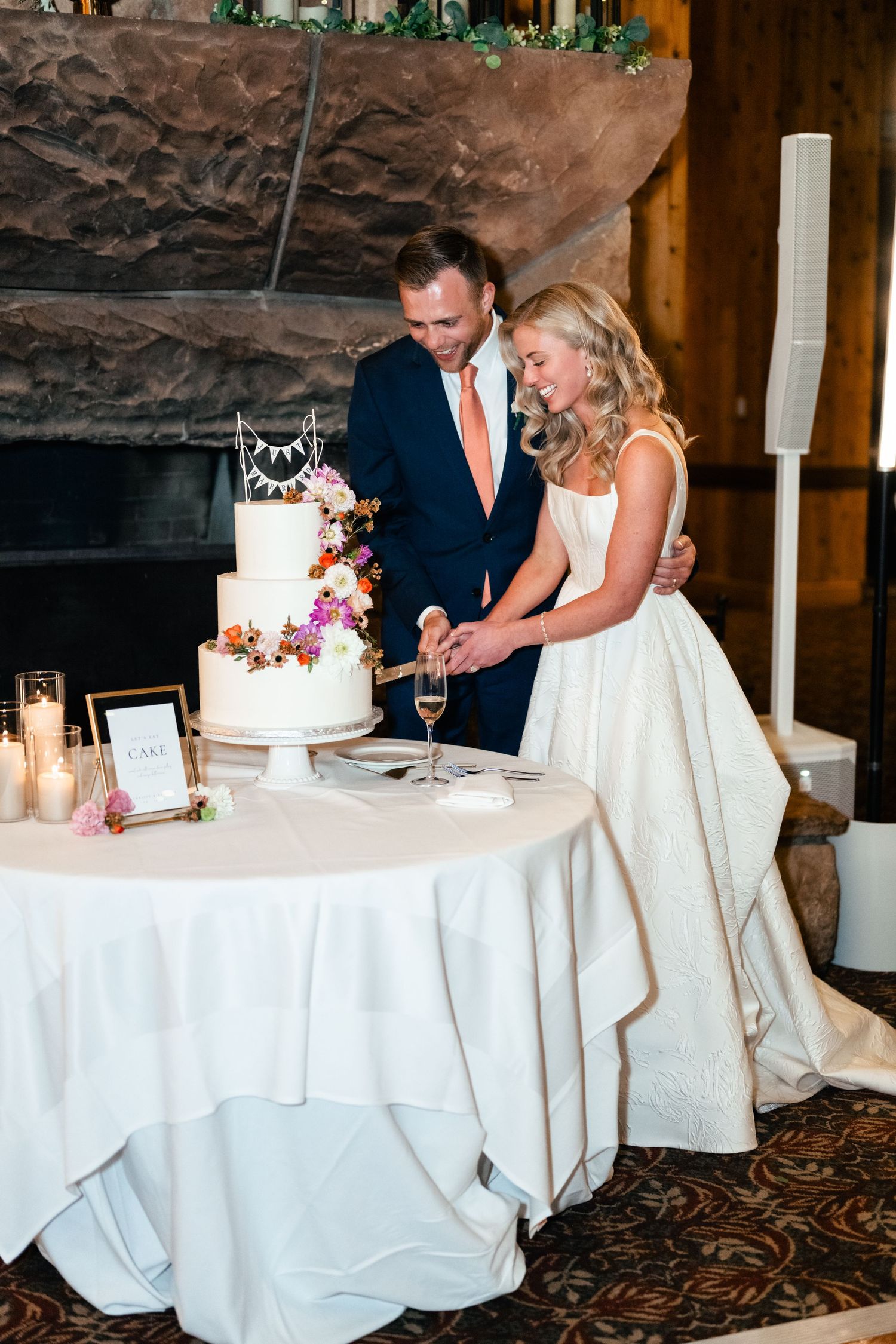 Wedding couple cutting their elegant white and floral decorated cake beside a stone fireplace.