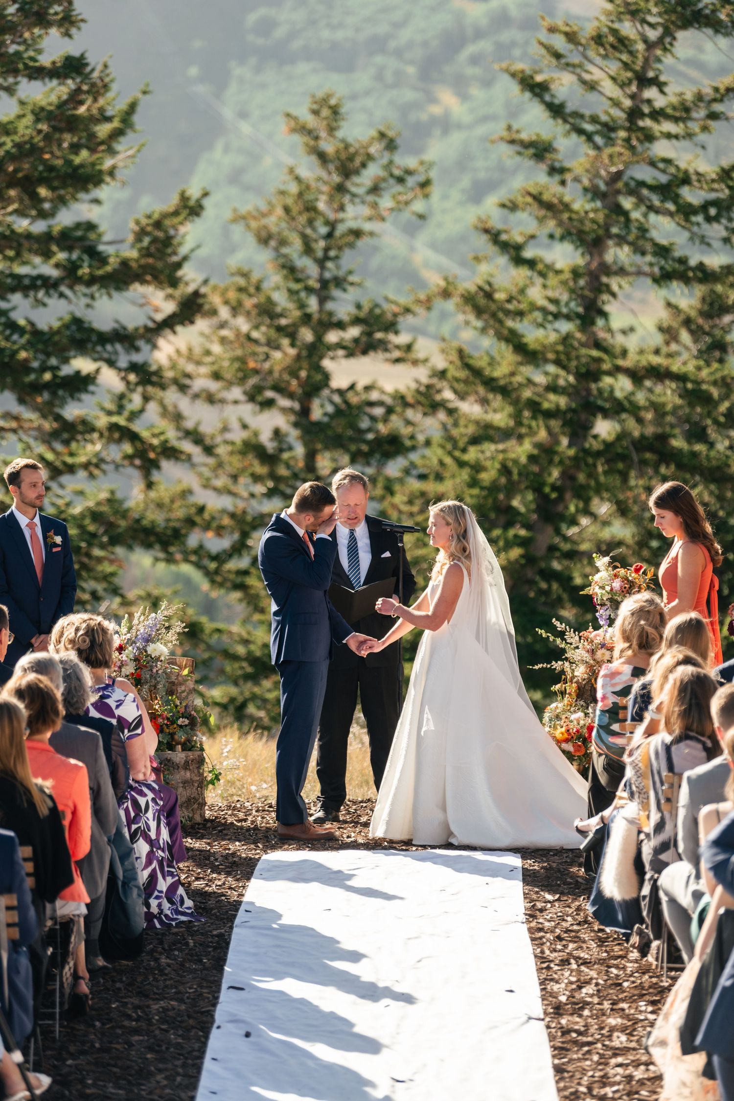 Outdoor mountain wedding ceremony with white aisle runner and evergreen trees in background.