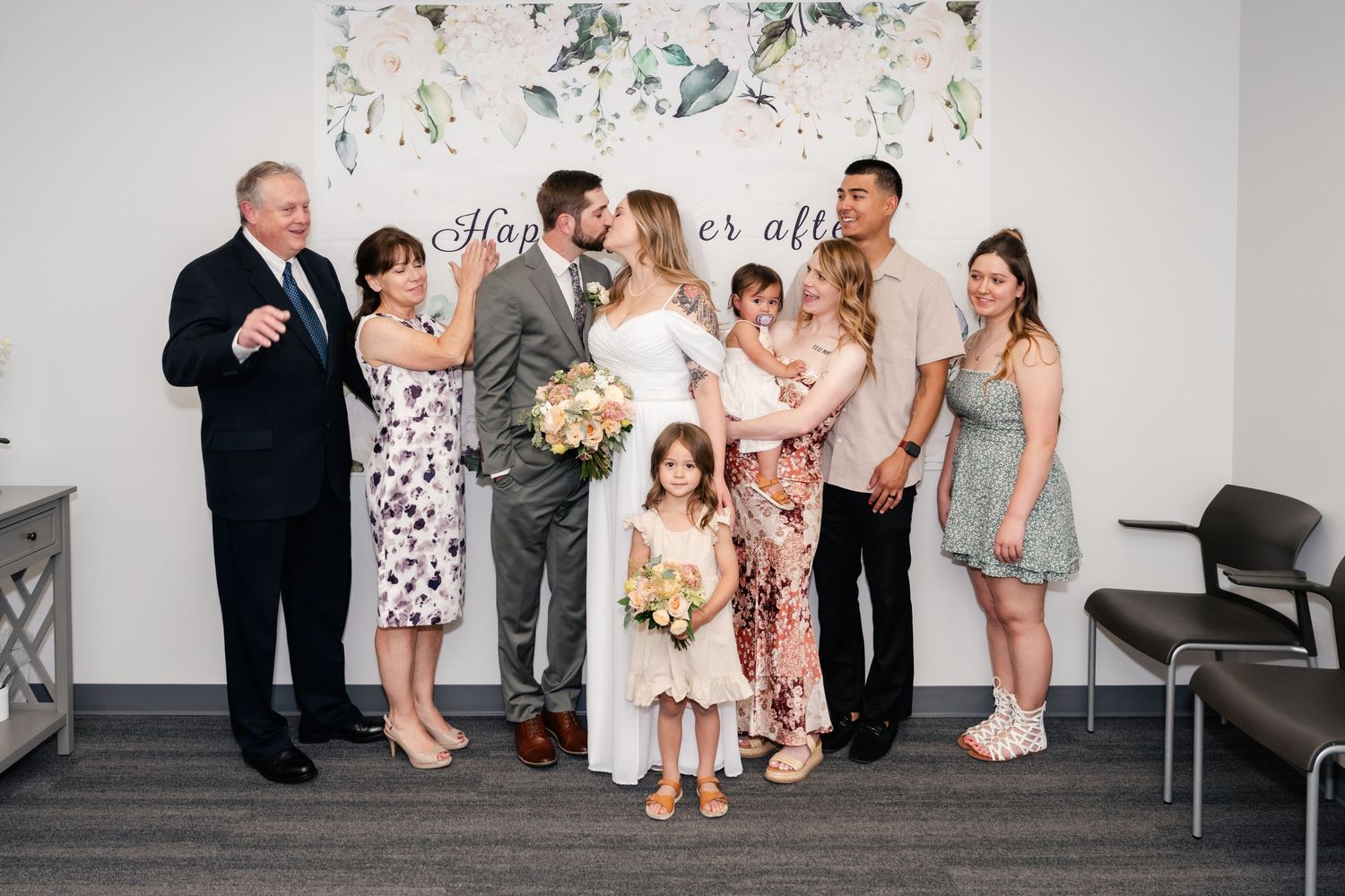 Wedding party poses for candid photos in a modern white room with floral decorations and a flower wall backdrop.