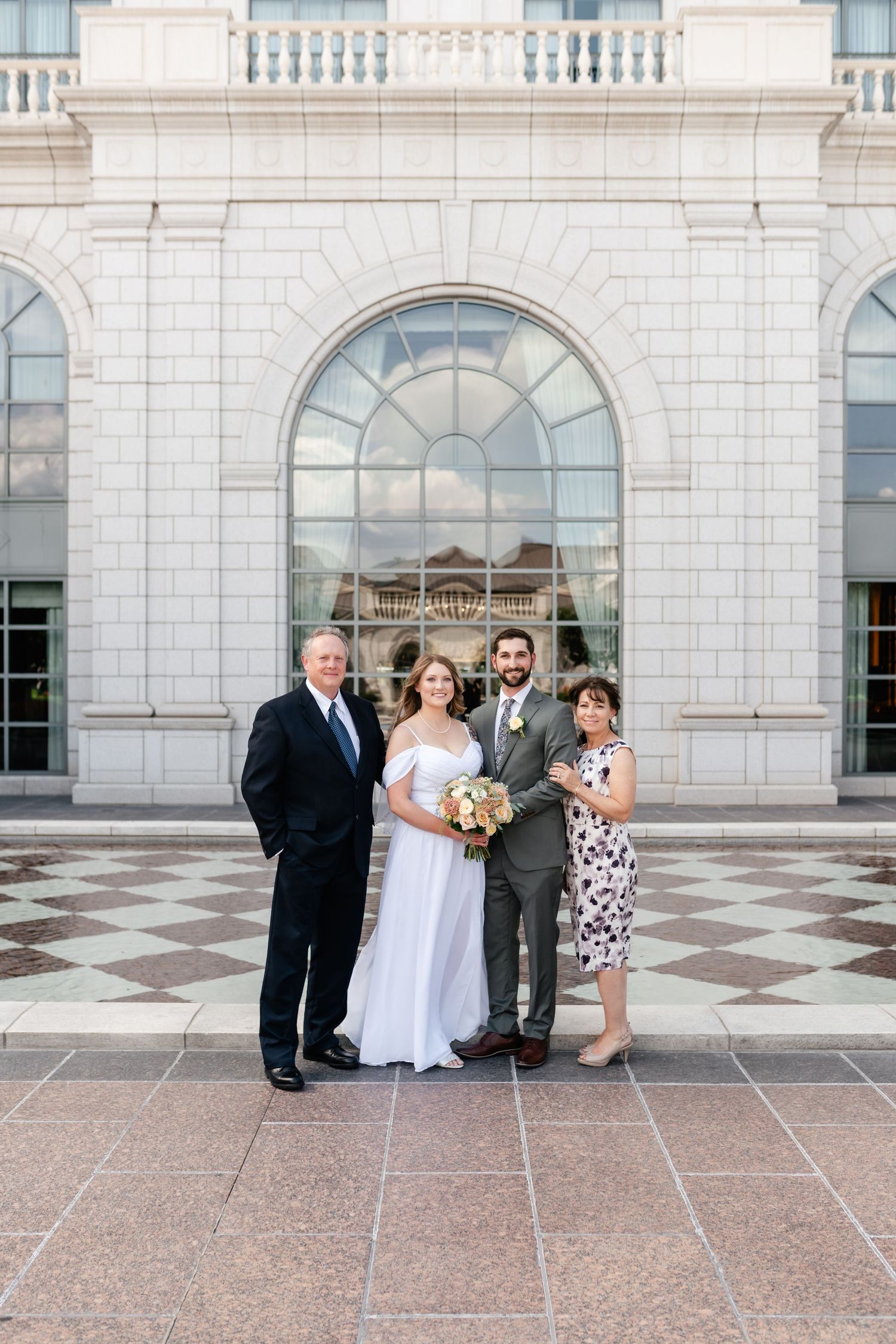 Wedding party poses for formal portraits on a checkered marble floor outside an elegant building.