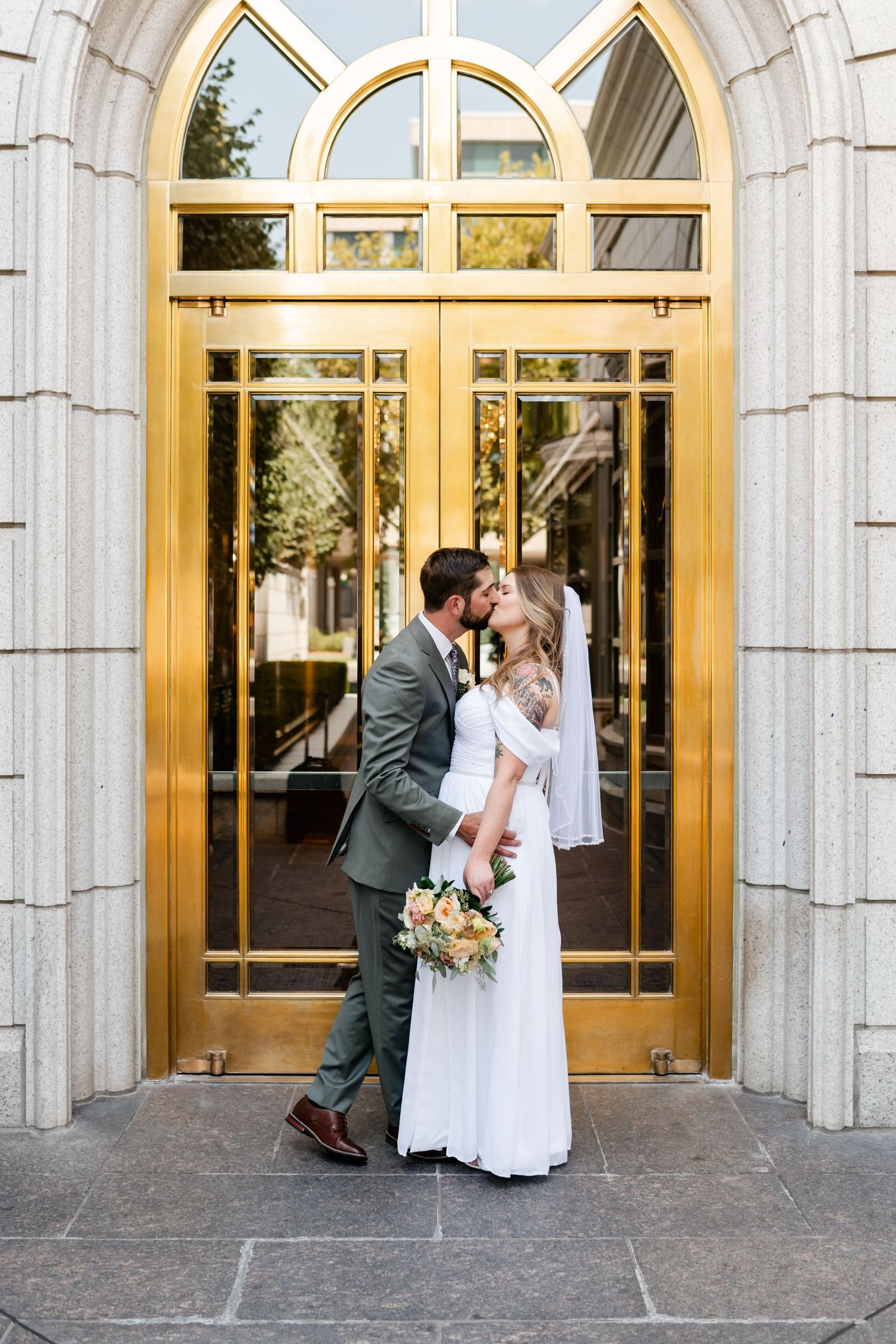 A romantic wedding couple shares a tender moment in front of grand arched church doors.