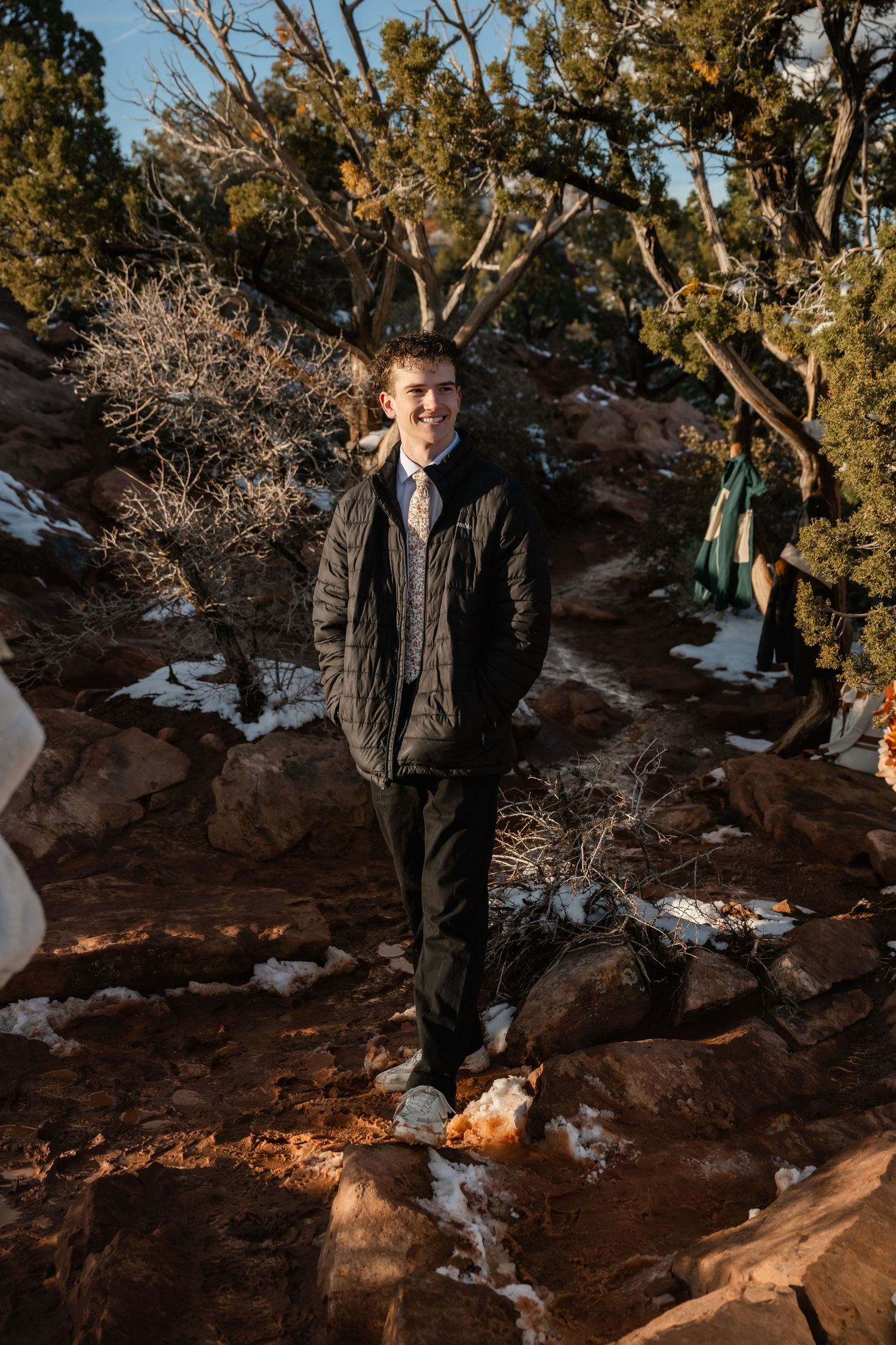 Figure in dark clothing stands on snowy red rocks with winter trees in background.