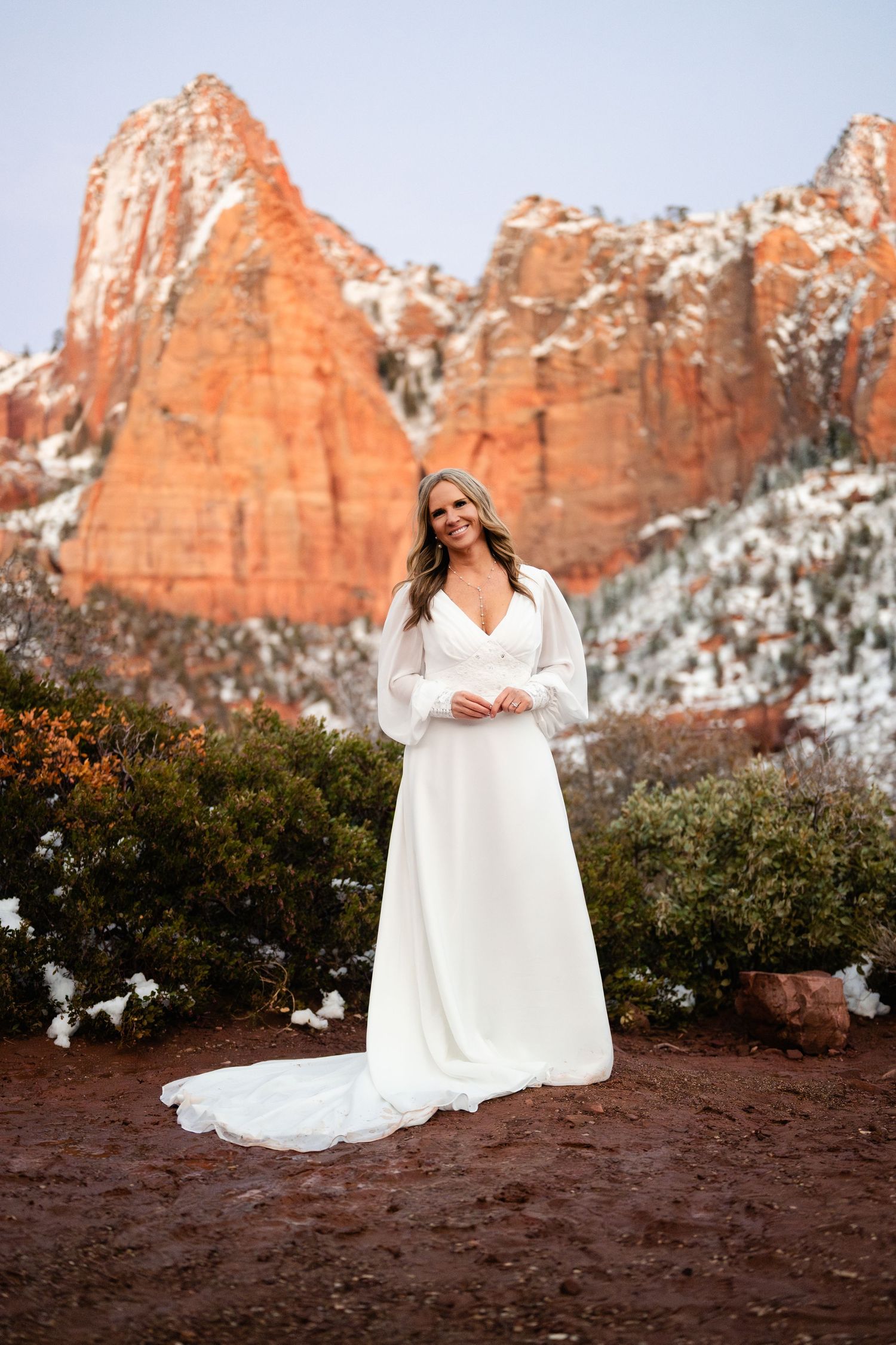Bride in white dress posing against snowy red rock cliffs during golden hour.