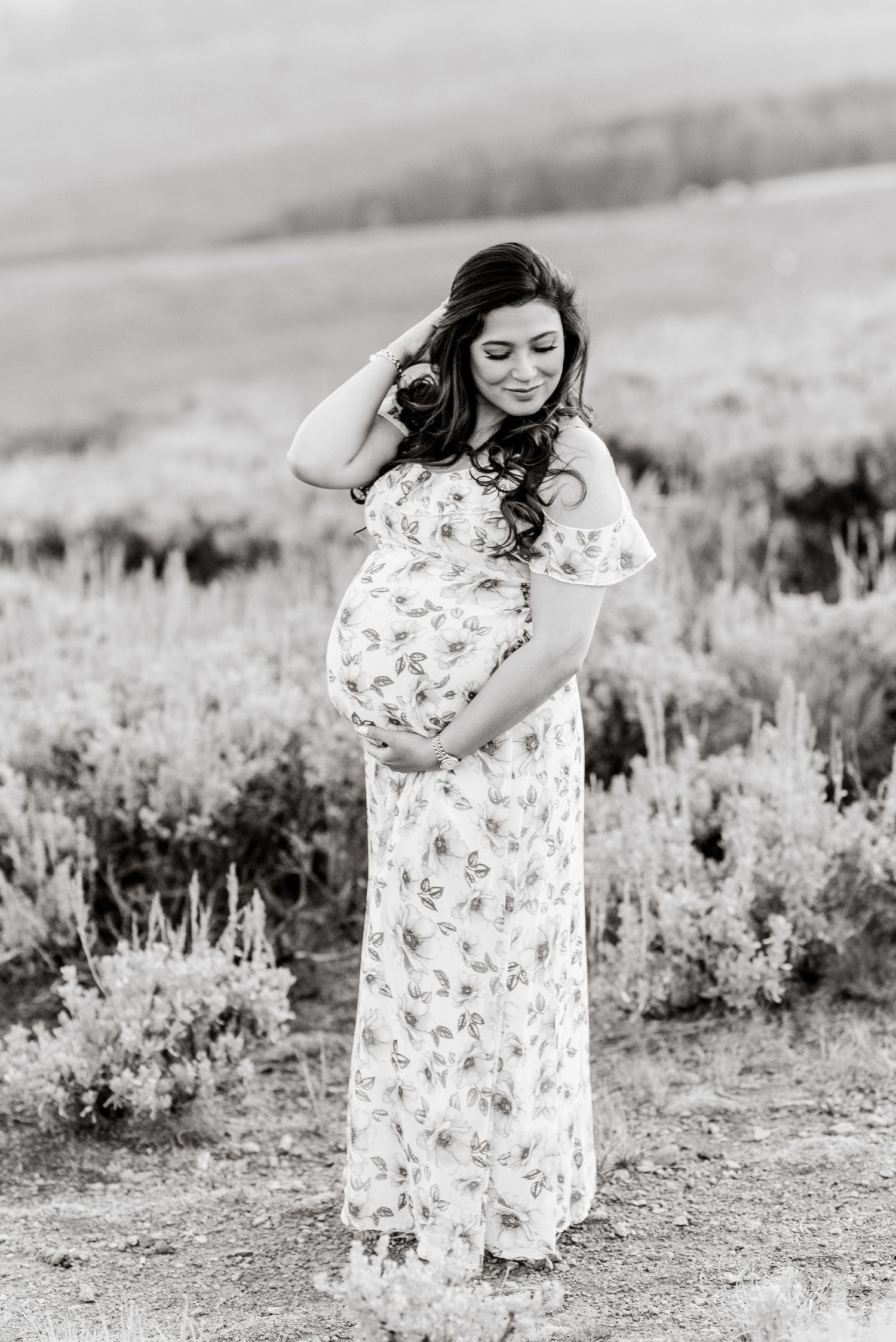 Black and white portrait of a woman in a floral dress standing amid wild sagebrush in a desert setting.