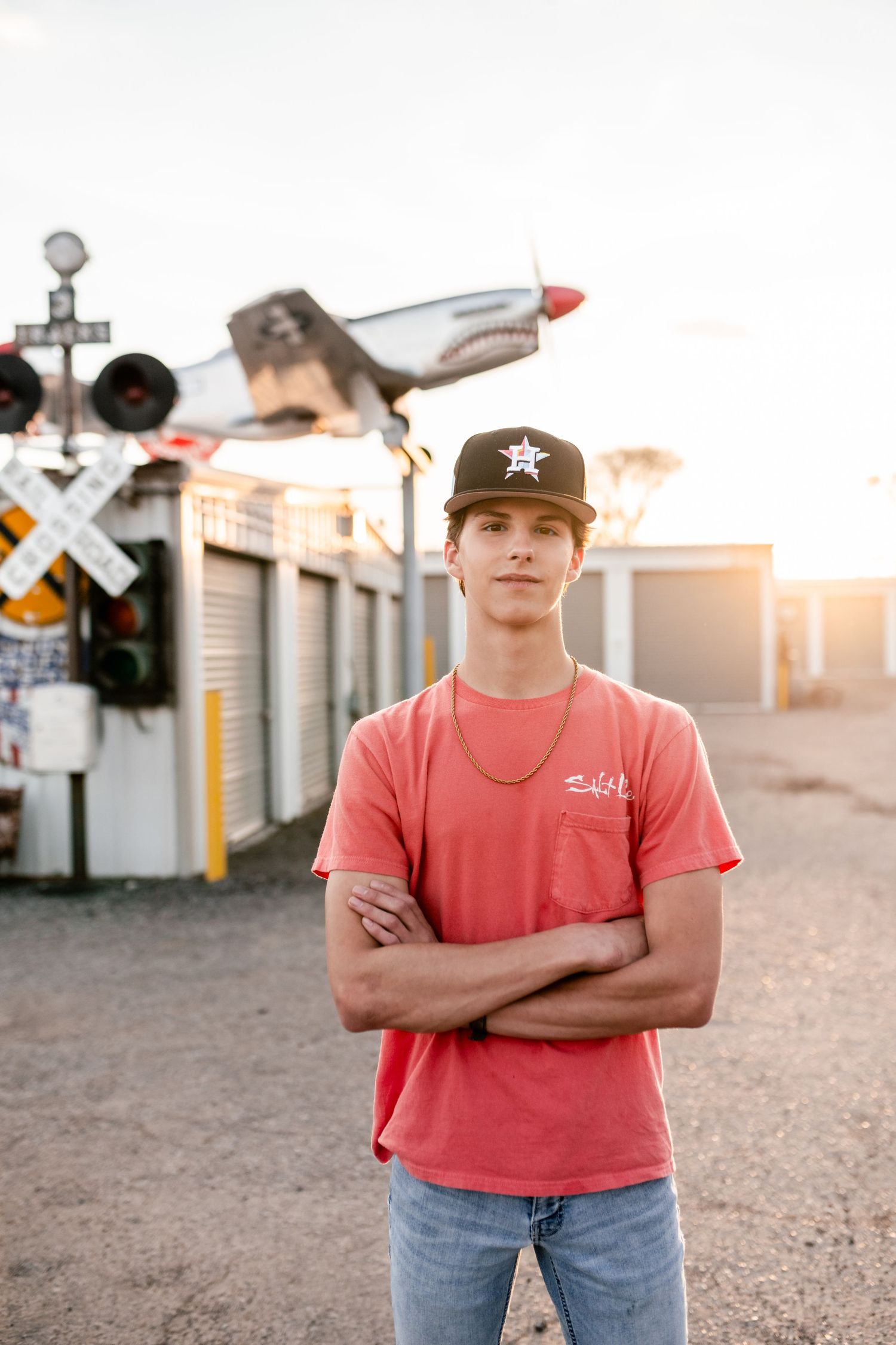 Person in a coral t-shirt and baseball cap stands near railroad tracks at sunset with industrial buildings visible.