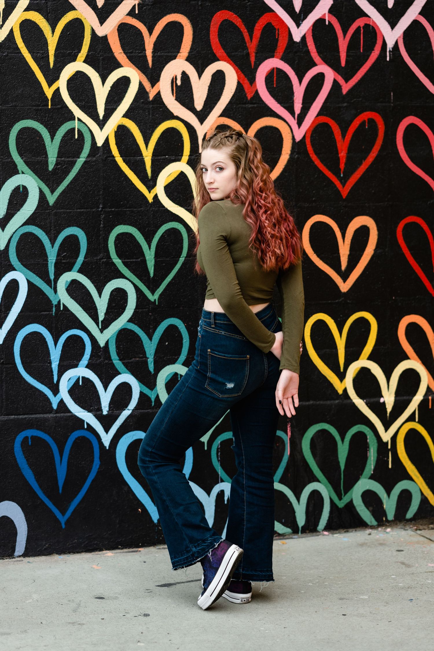 A person poses against a vibrant wall mural featuring colorful hearts painted in rainbow hues on a black background.