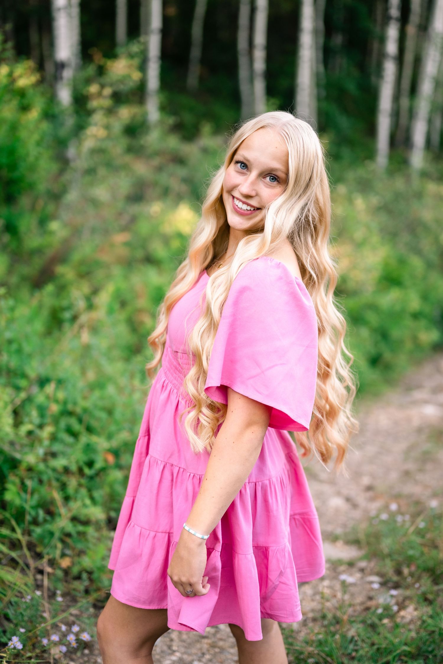 A person in a pink babydoll dress poses on a forest path with long wavy blonde hair and a bright smile.