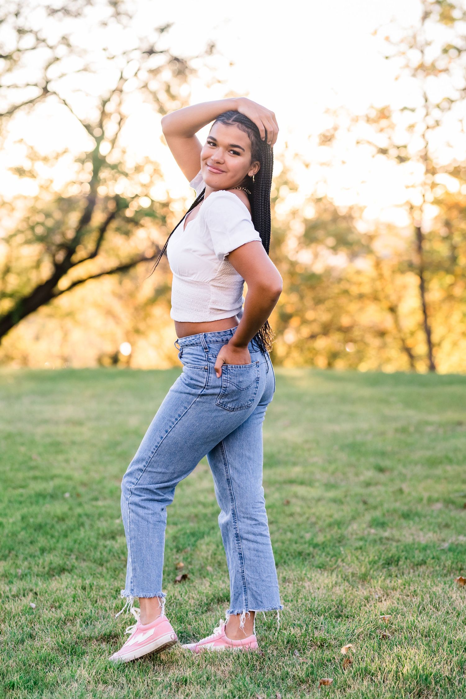 Person in casual outfit of white crop top and blue jeans poses playfully on grassy field at sunset.