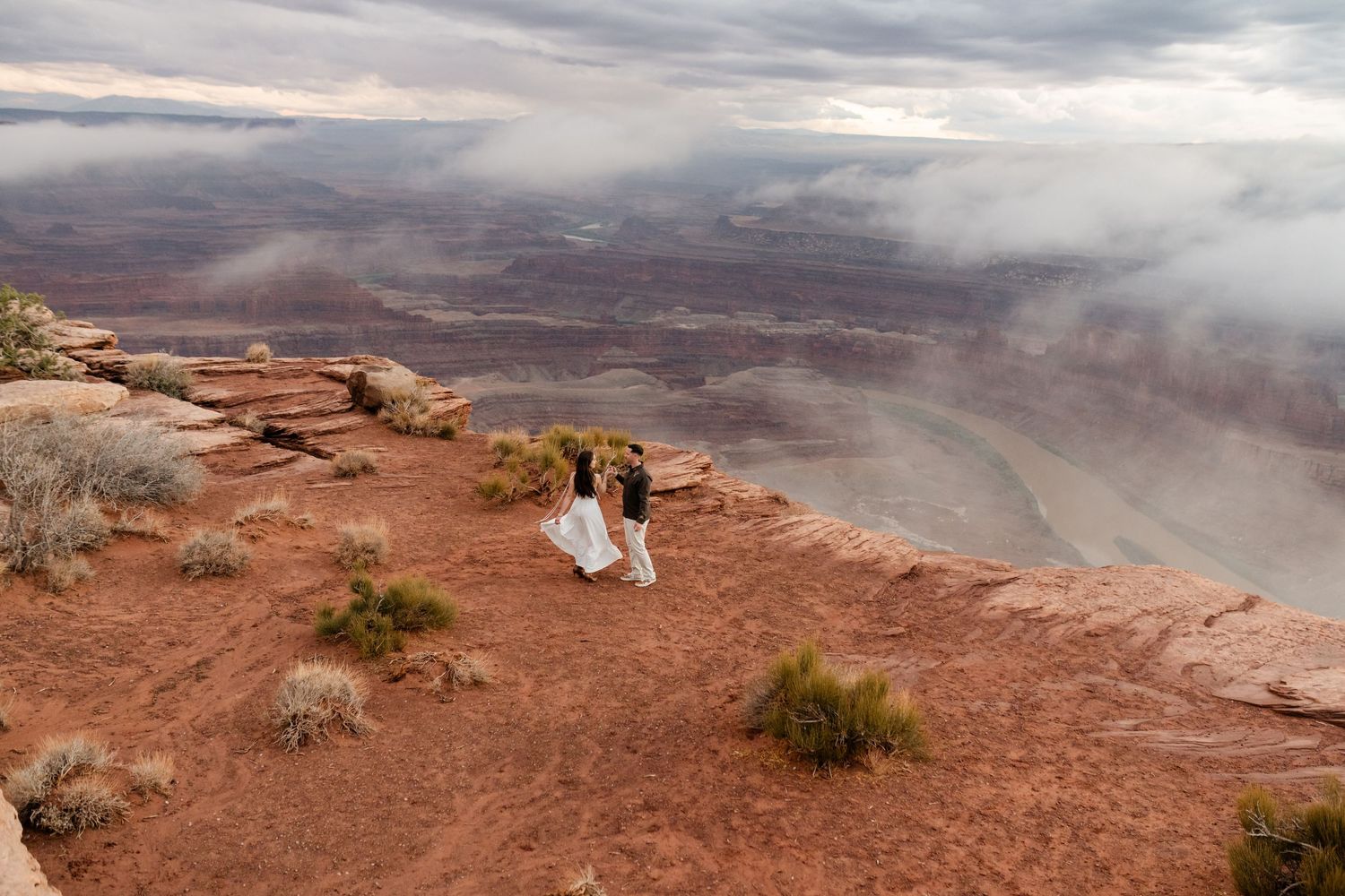 A couple stands on red desert cliffs overlooking a misty canyon landscape.