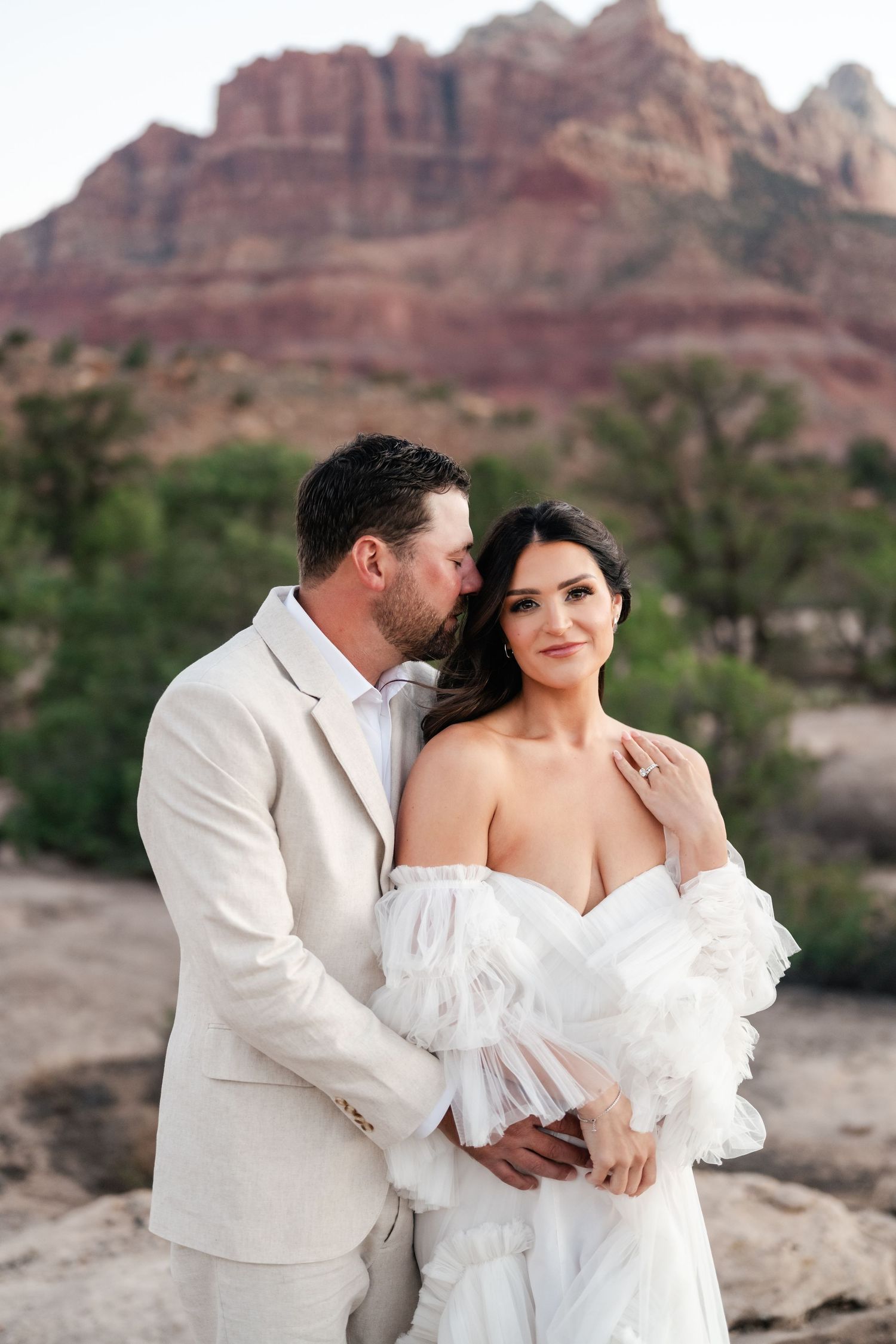 Zion destination photographer. A romantic couple in white attire embraces against a backdrop of red rock formations in the desert.