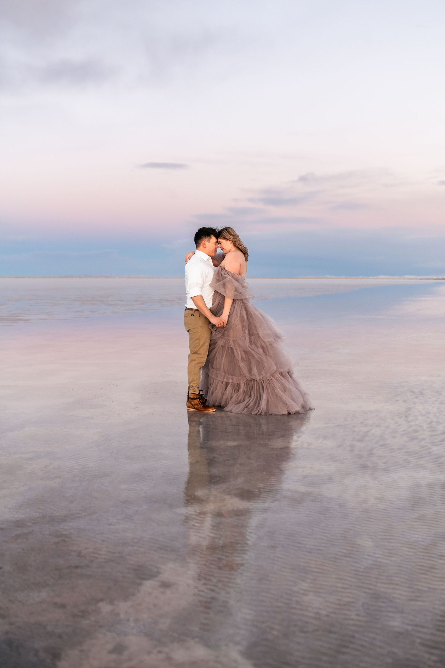 Utah Salt Flats Photographer. A couple embraces on a reflective salt flat during sunset with soft pink and blue sky.