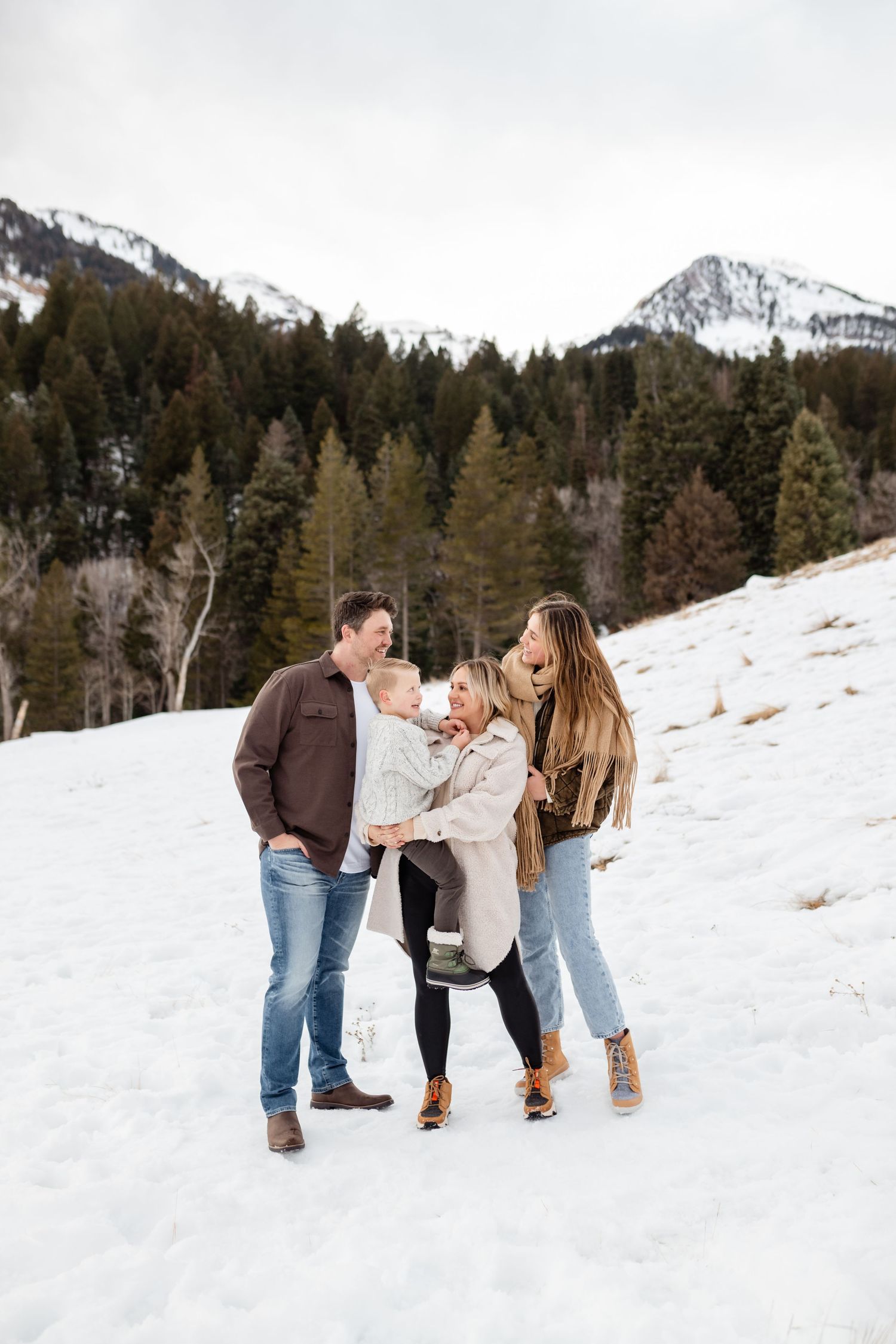 Family of three playing together in the snow with mountain peaks and evergreen trees in the background.
