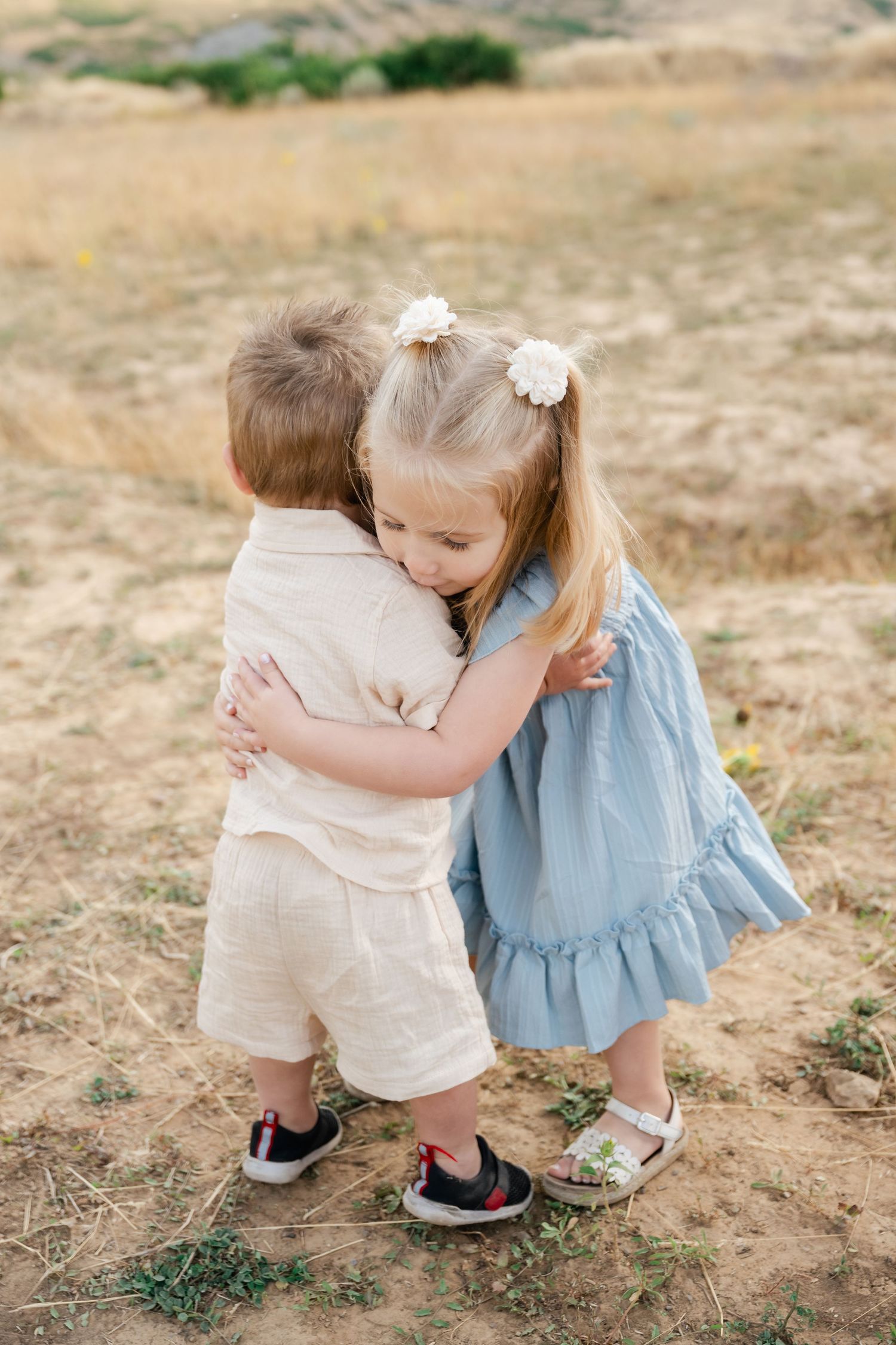 Two young children share a sweet hug while standing on a dirt path outdoors.