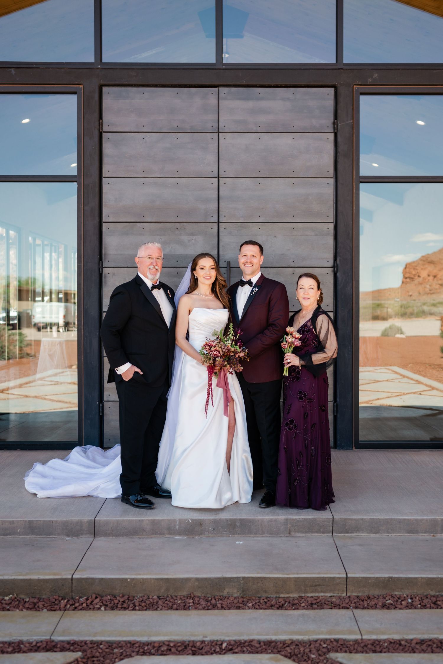 Wedding party of four poses outside modern venue, bride in white gown with groom in burgundy tuxedo flanked by family.