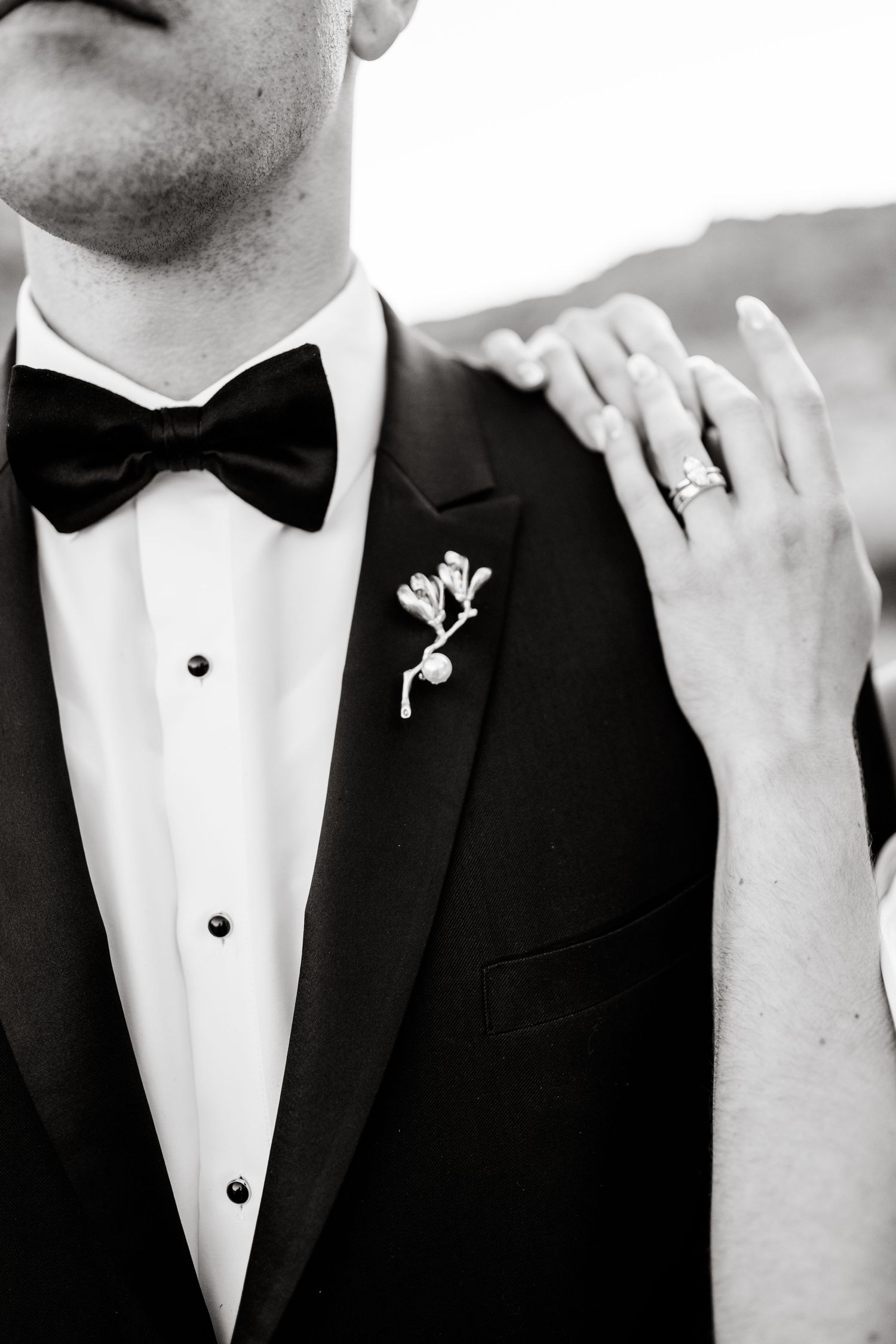 Black and white close-up of groom in tuxedo with bow tie and floral boutonniere, bride rests hand on his shoulder showing ring.