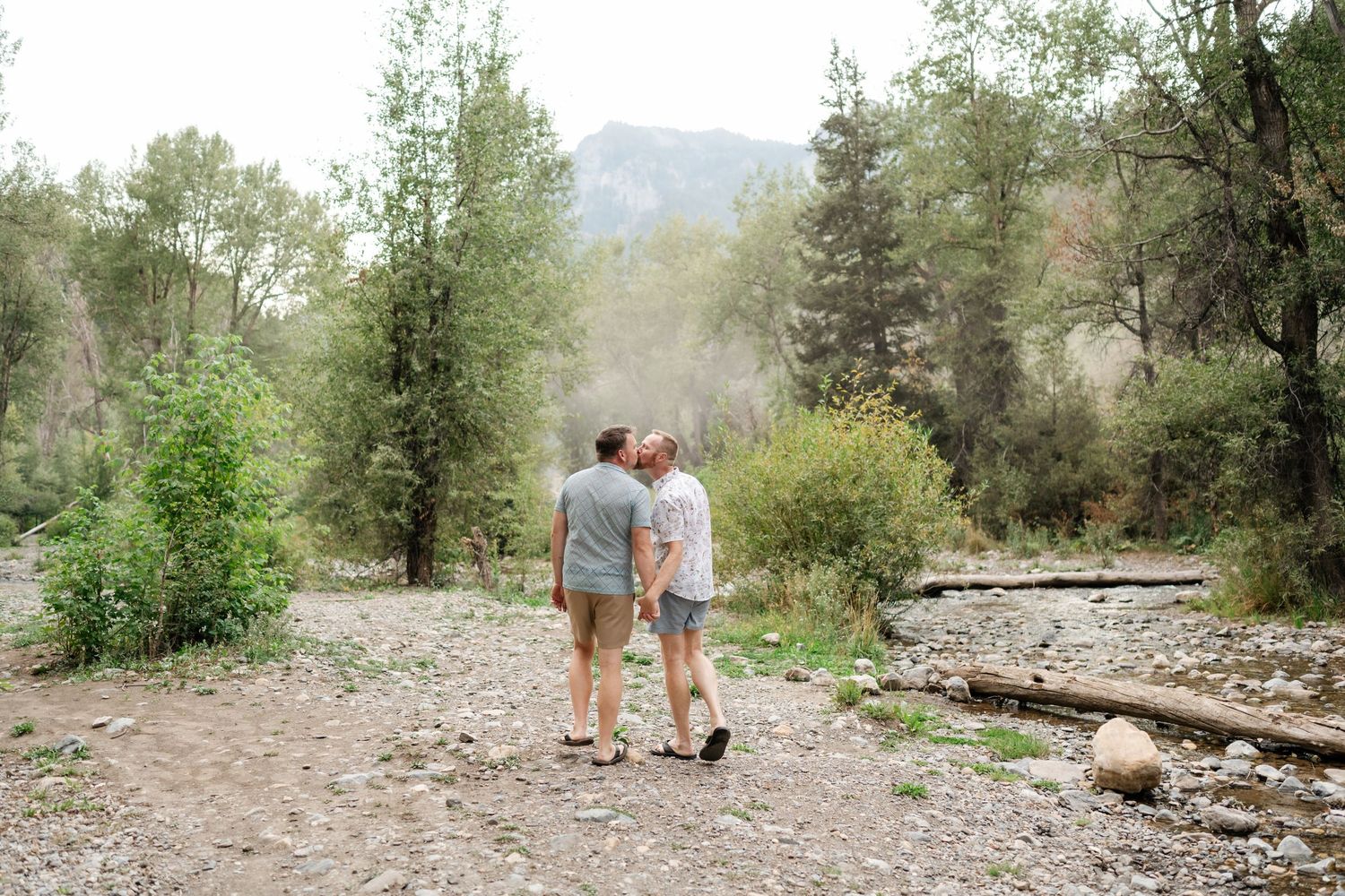LBTQ couple walk hand-in-hand along a rocky mountain path surrounded by lush green trees and misty hills in American Fork Canyon.