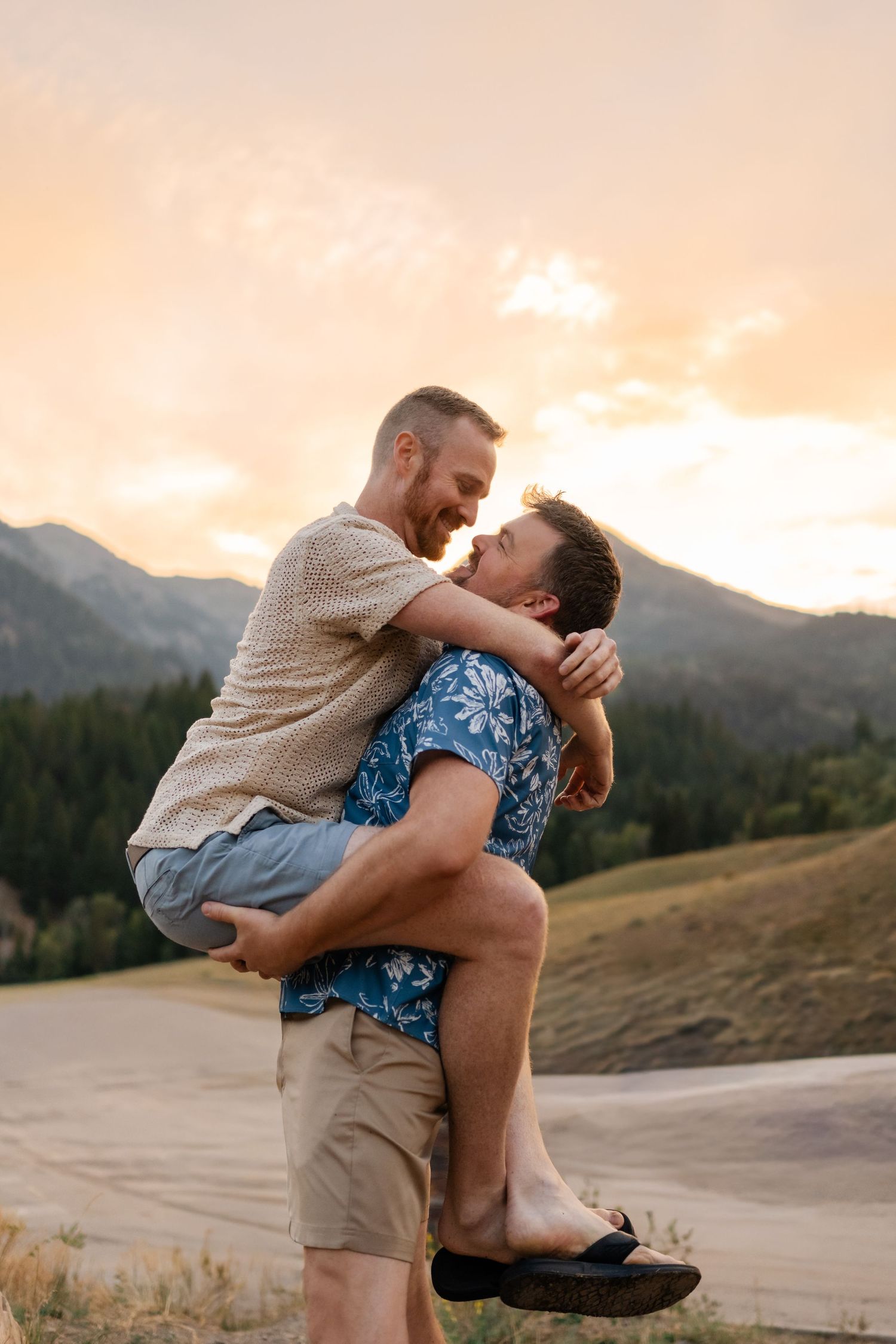 Two men share a romantic embrace at sunset with mountain scenery backdrop, one lifting the other joyfully.
