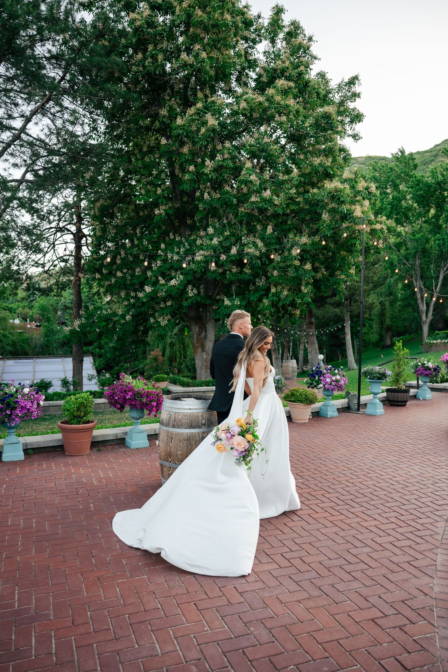 A couple in wedding attire shares a romantic spin on a red brick patio surrounded by lush garden landscaping.