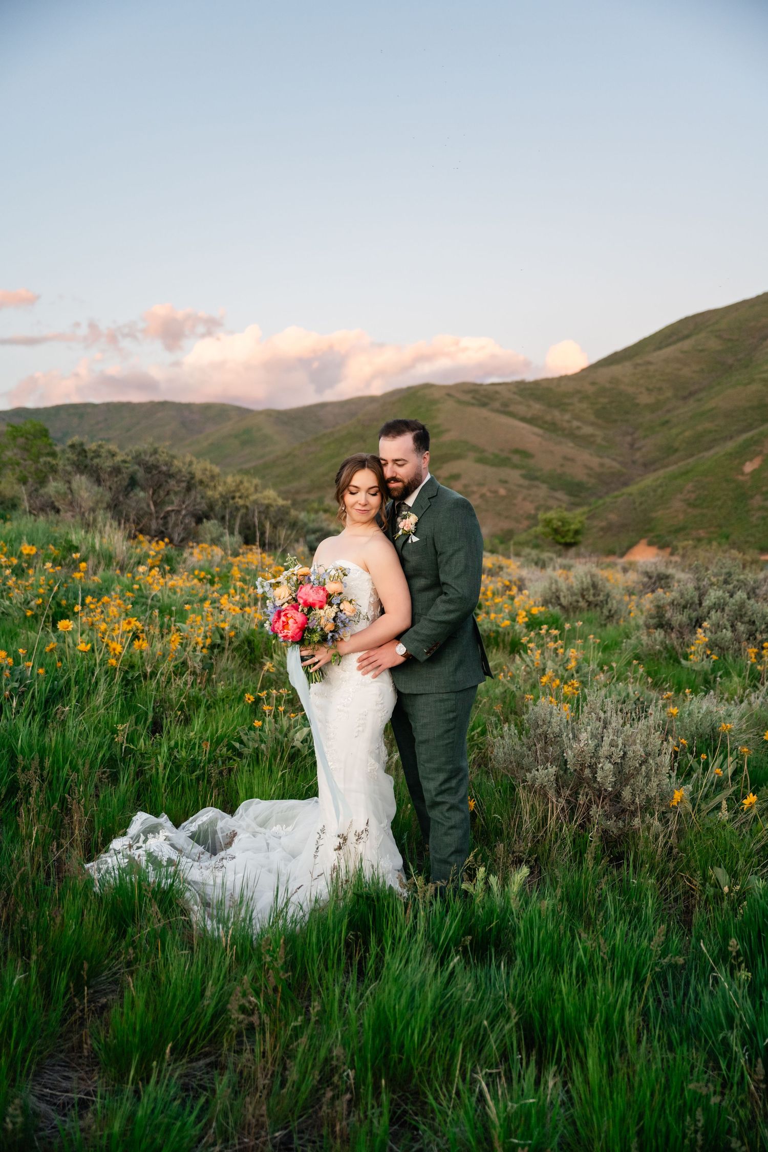 A romantic wedding couple stands among colorful wildflowers in a mountainous landscape at sunset.
